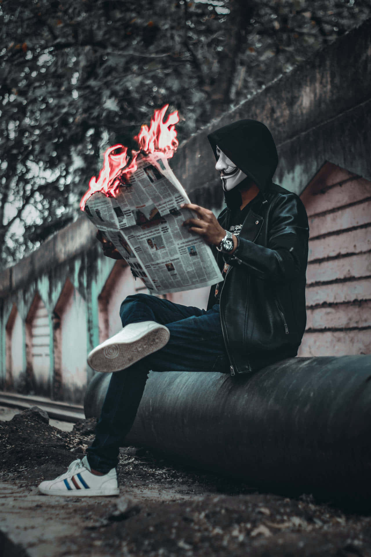 Mask 桌布 壁紙 — 一個戴著面具、身穿黑色連帽衫的人物坐在管道上，在 gritty 的城市背景下閱讀燃燒的報紙。
