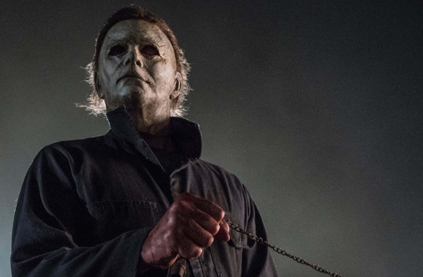 Mike Myers 桌布 壁紙 — 一個戴著面具的威脅人物，身穿深色外套，在陰影背景中拿著一條鏈條。
