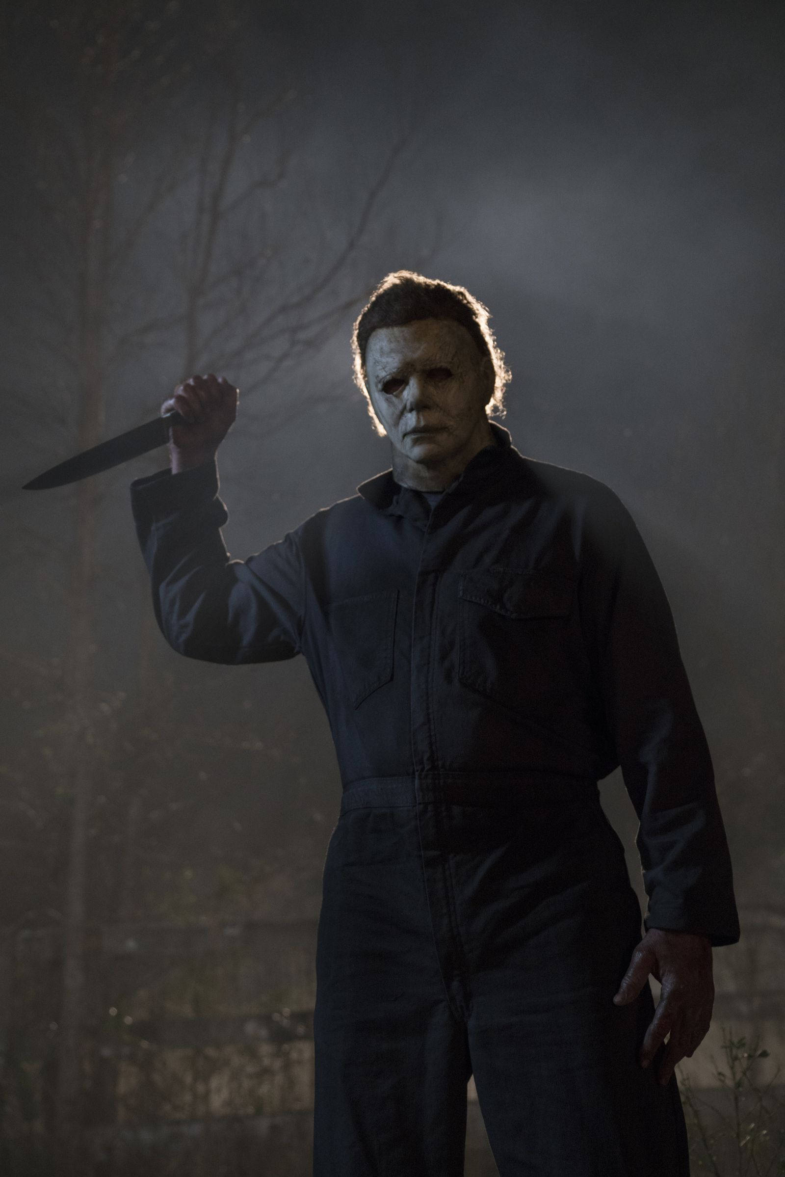 Halloween Kills 桌布 壁紙 — 一個戴著面具的威脅人物身穿深色連身工作服，在霧氣籠罩的樹林區域中，在戲劇性的背光下拿著一把刀。