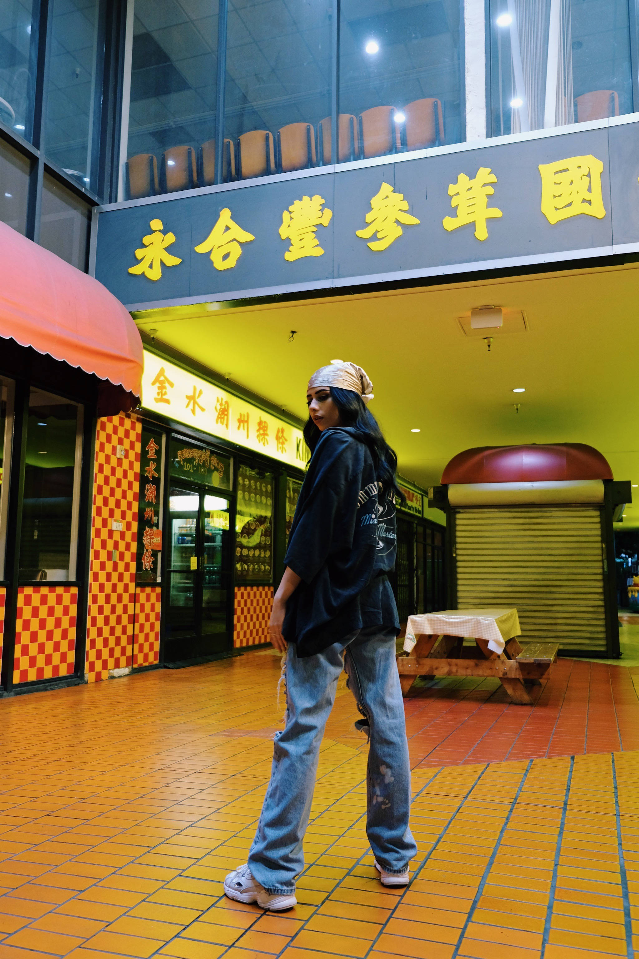Tomboy 桌布 壁紙 — 一個戴著頭巾和超大外套的人走過一個光線充足的室內美食廣場，地板鋪有黃色瓷磚，並有中文標誌。