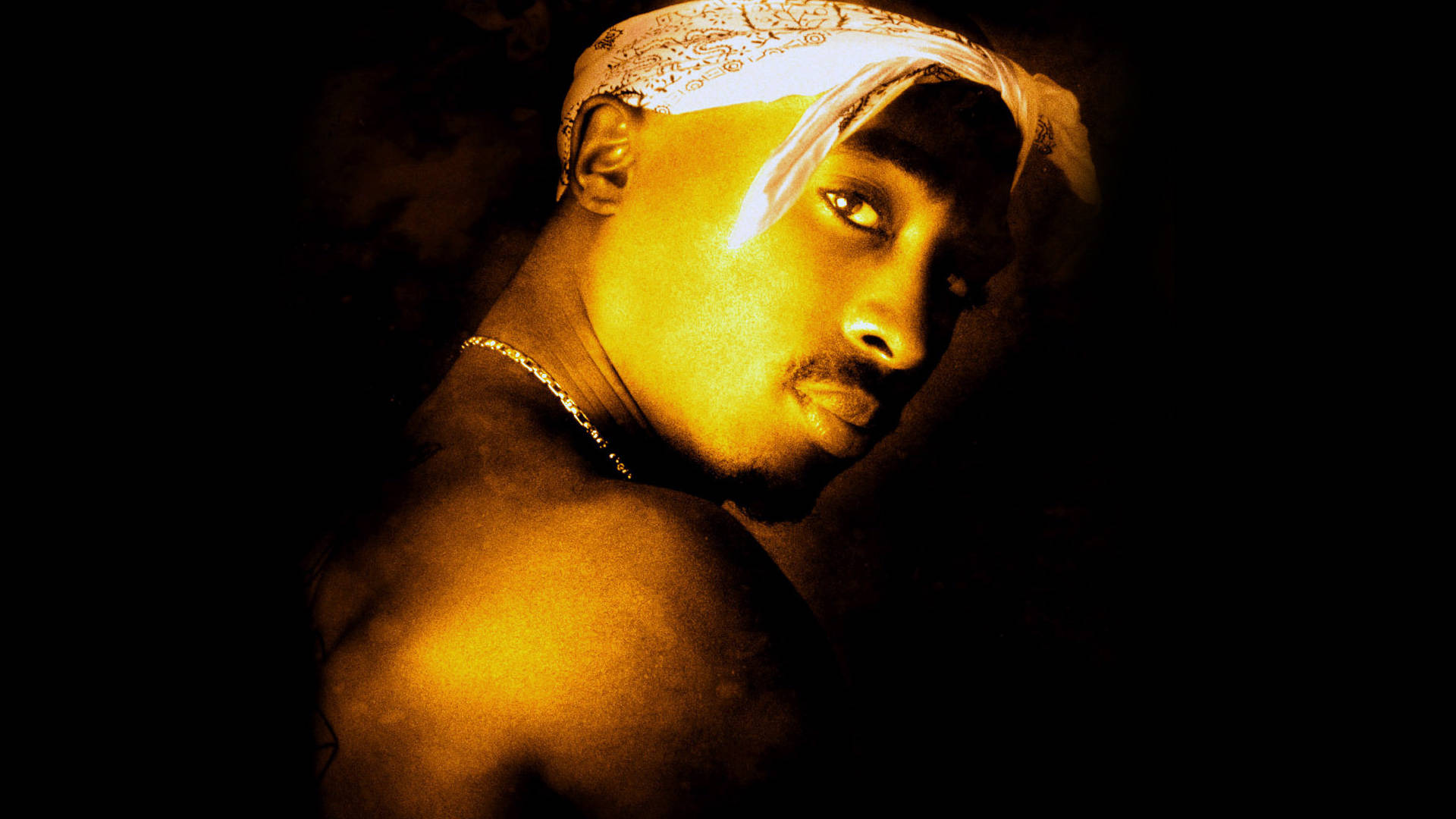 tupac 桌布 壁紙 — 一個戴著頭巾和鏈條的男人在黑暗背景下閃耀著金色光芒。