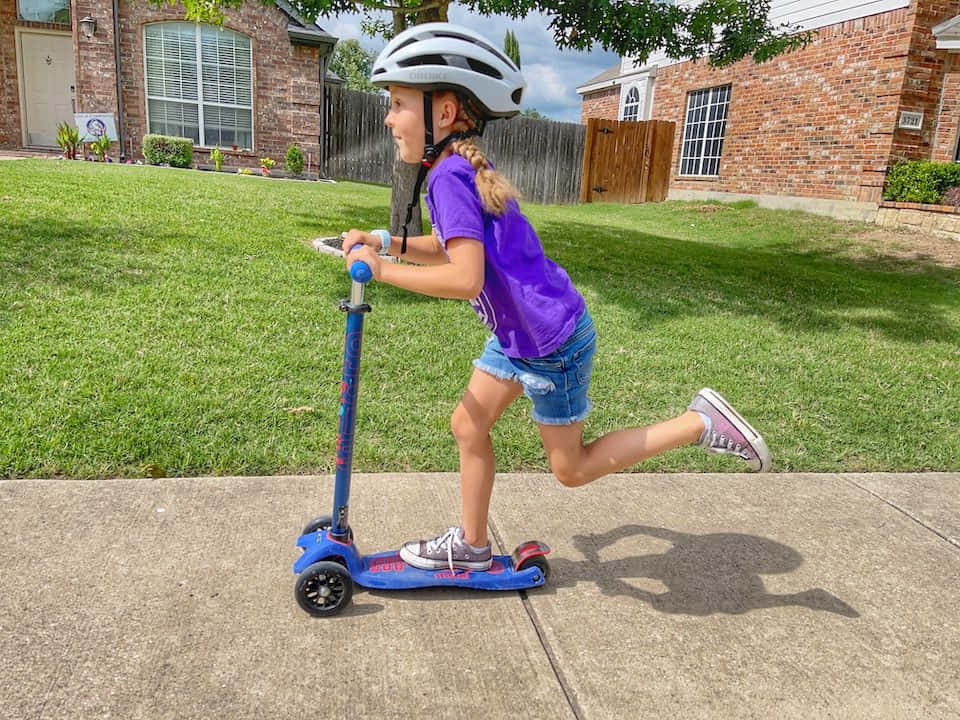 scooter 桌布 壁紙 — 一個戴著頭盔、穿著紫色襯衫的小女孩，在陽光明媚的郊區後院混凝土小路上騎著藍色滑板車。