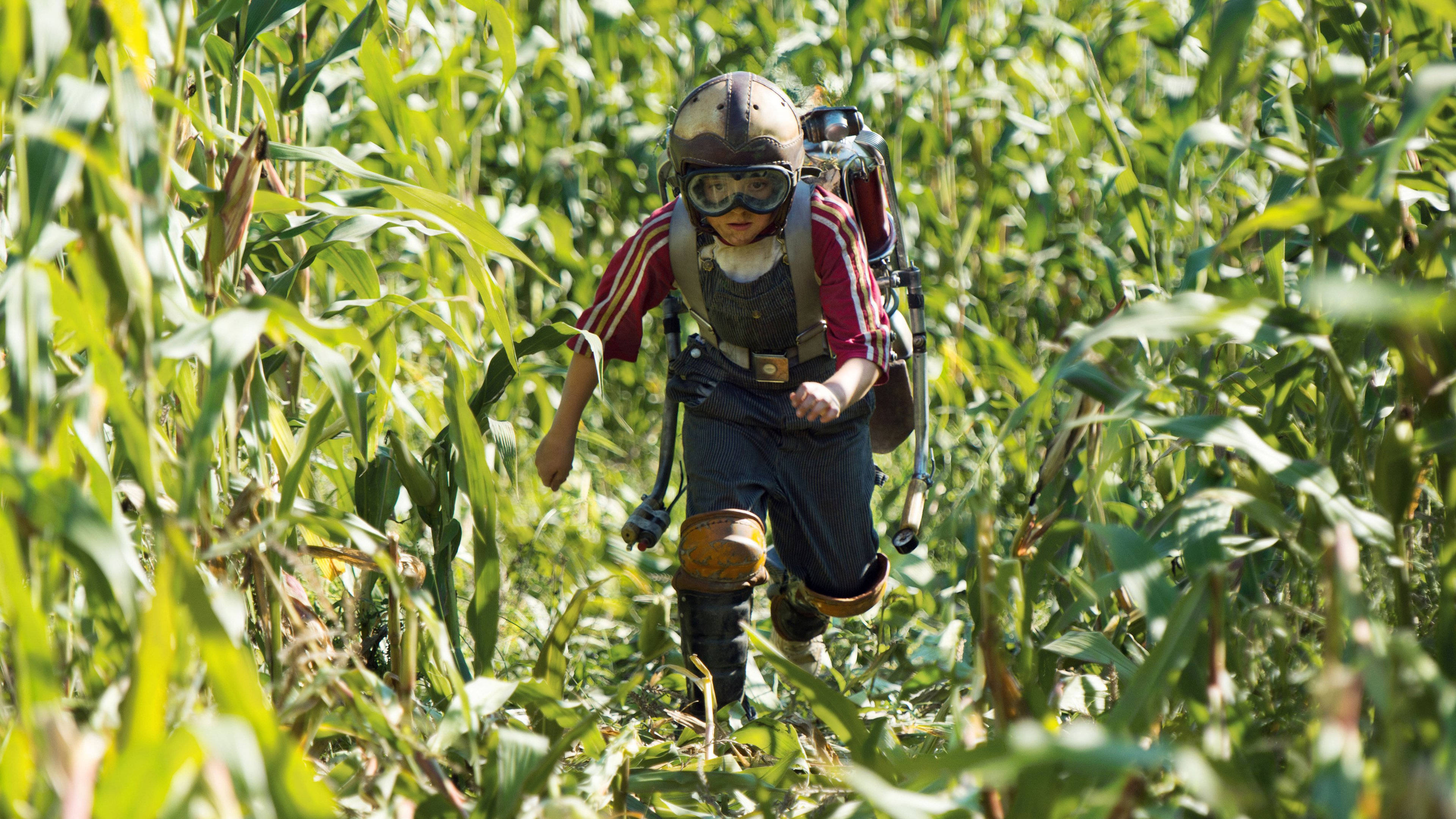 Tomorrowland Movie 桌布 壁紙 — 一個戴著頭盔和護目鏡的孩子背著背包降落傘在陽光明媚的玉米田裡奔跑。