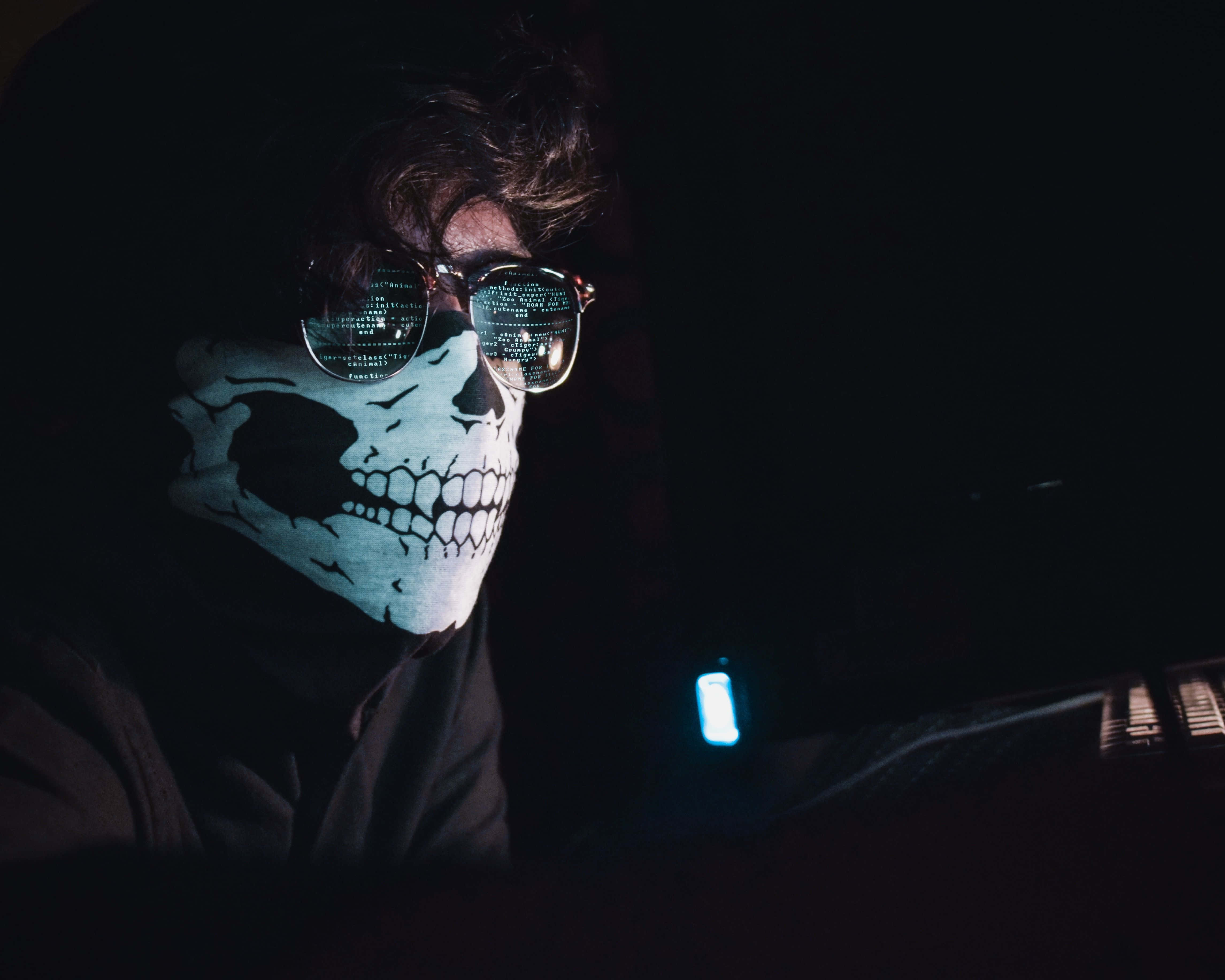 Horror Masks 桌布 壁紙 — 一個戴著骷髏面具和眼鏡的人，眼鏡反射著數位代碼，坐在黑暗中，被發光的藍色螢幕照亮。