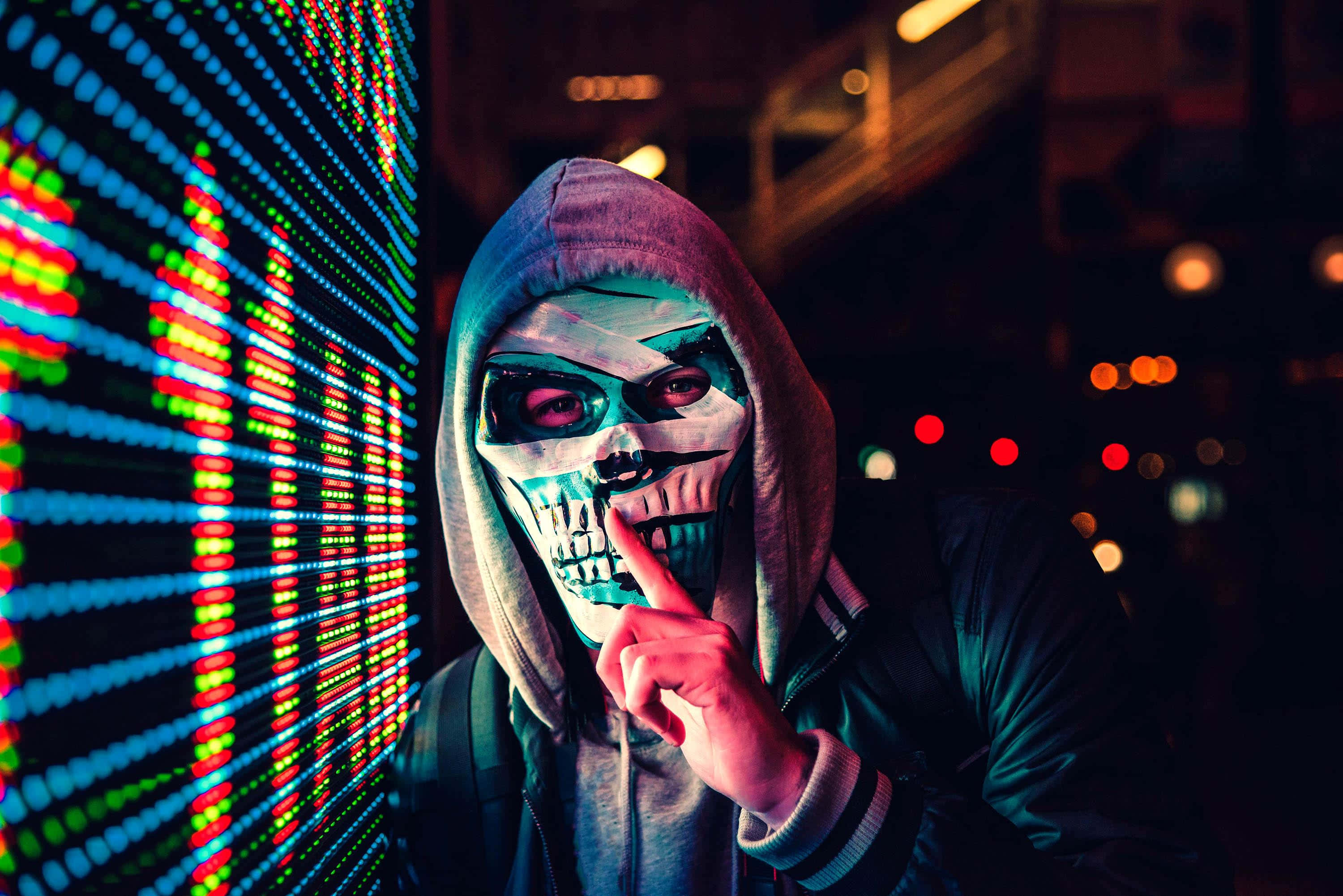 Mask 桌布 壁紙 — 一個戴著骷髏面具和連帽衫的人，在黑暗的城市夜景中，旁邊有一個充滿活力的LED顯示屏，將手指放在嘴唇上。