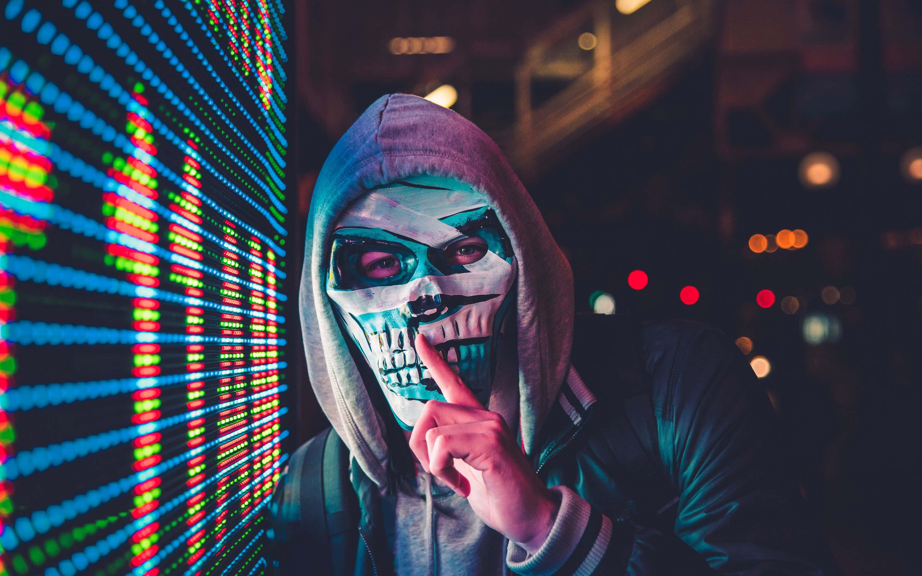 Horror Masks 桌布 壁紙 — 一個戴著骷髏面具和連帽衫的人站在黑暗城市夜晚環境中發光的數位螢幕前。