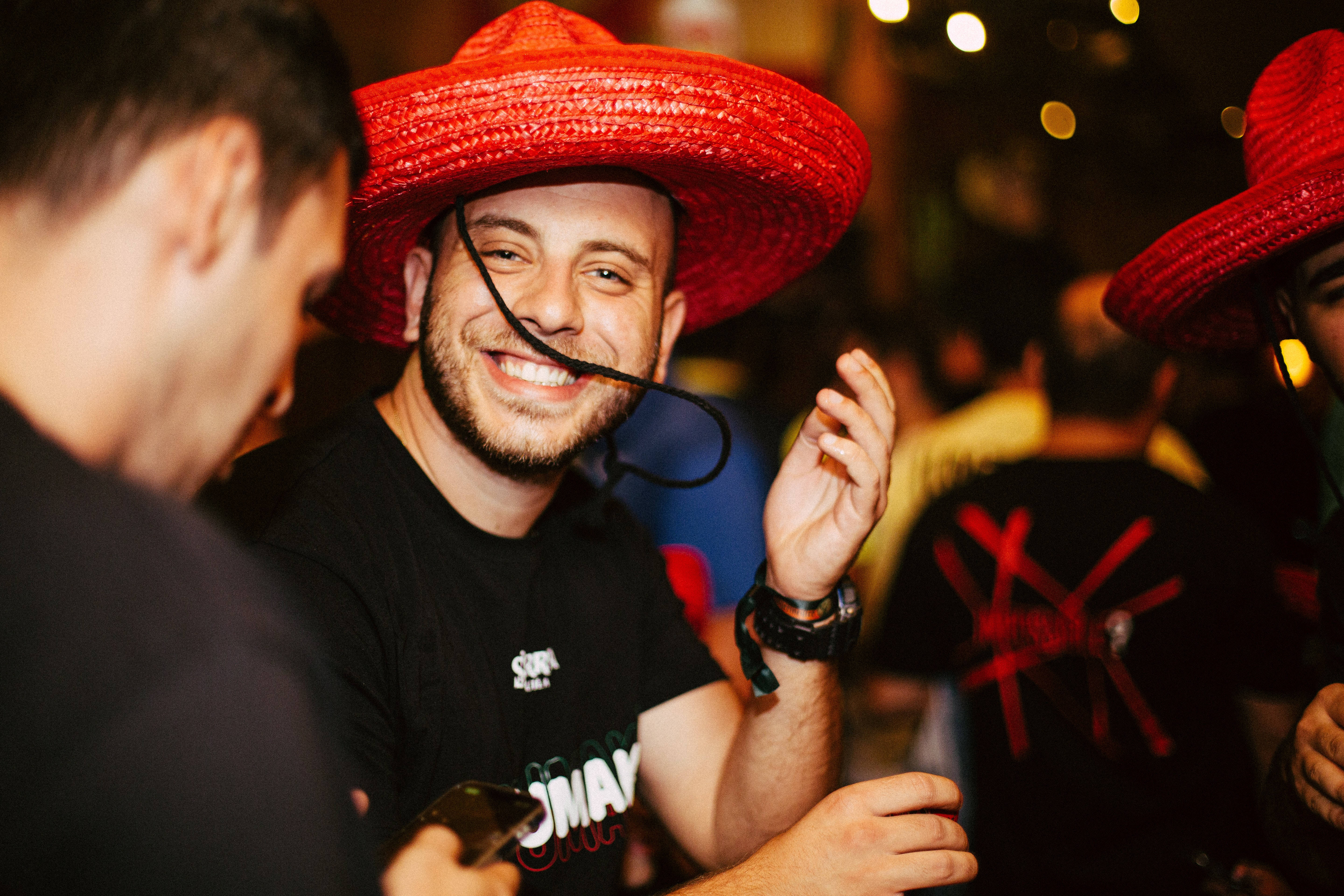 Mexican 桌布 壁紙 — 一個戴著鮮紅色闊邊帽的男人在熱鬧的夜間聚會中咧嘴大笑，同時將一條黑色繩子靠近嘴邊。