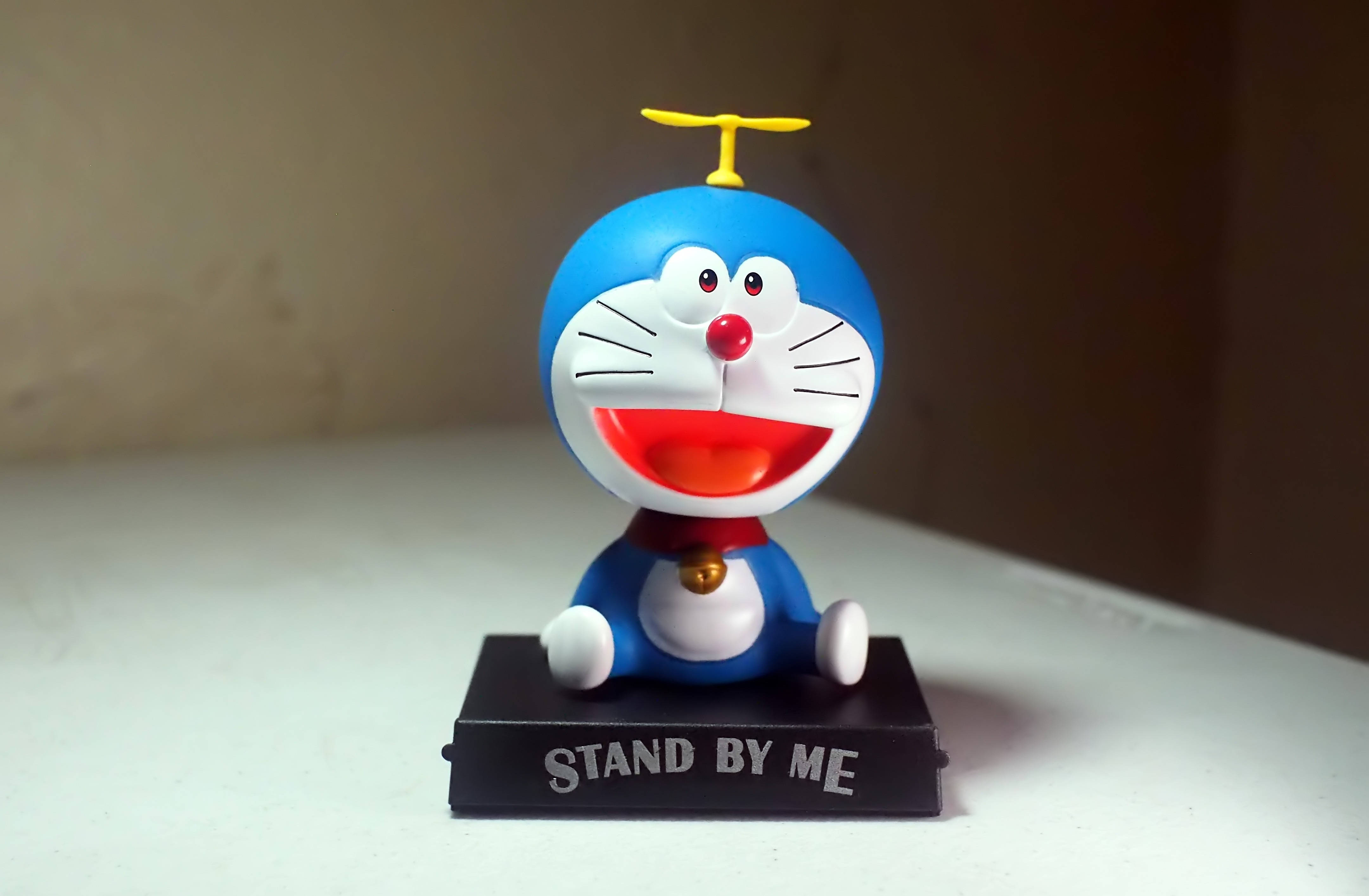 Doraemon 桌布 壁紙 — 一個戴著黃色螺旋槳帽的開心藍色機器貓公仔，坐在黑色底座上，底座上寫著「STAND BY ME」，放在白色表面上。
