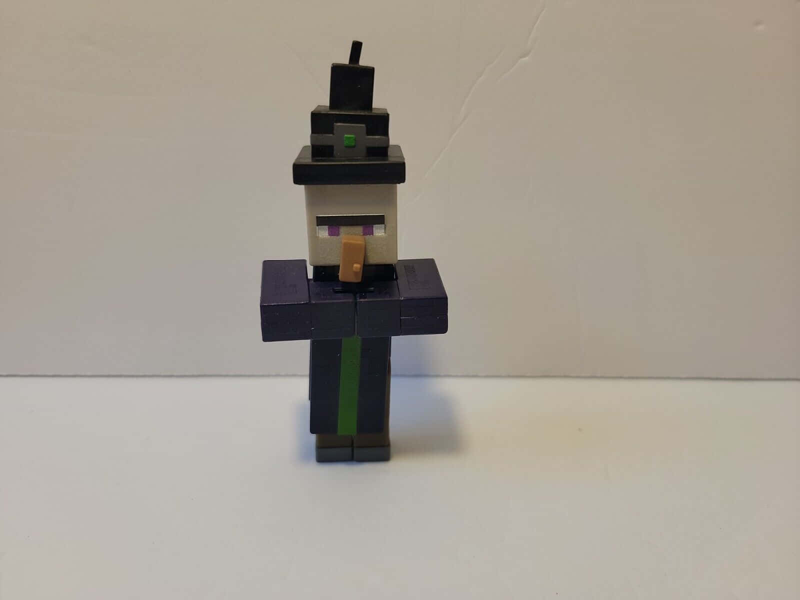 Minecraft Witch 桌布 壁紙 — 一個戴著黑色帽子、綠色眼睛和紫色長袍的 Minecraft 女巫公仔，站在純白色背景前。