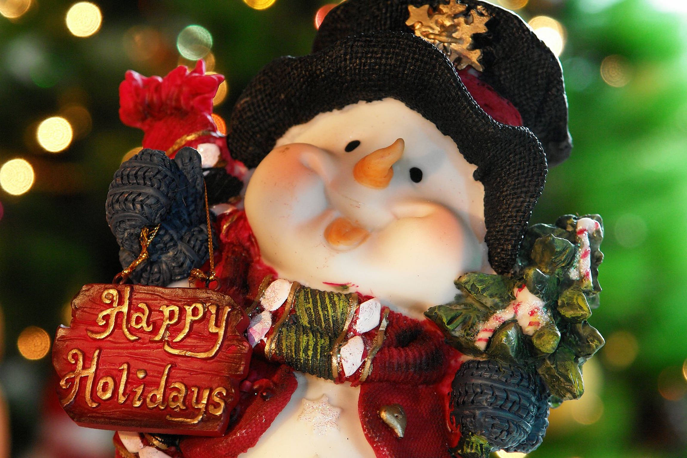 可愛聖誕節 桌布 壁紙 — 一個戴著黑色帽子和紅色圍巾的快樂雪人公仔，在綠色和金色燈光的散景背景前舉著「Happy Holidays」標語牌。