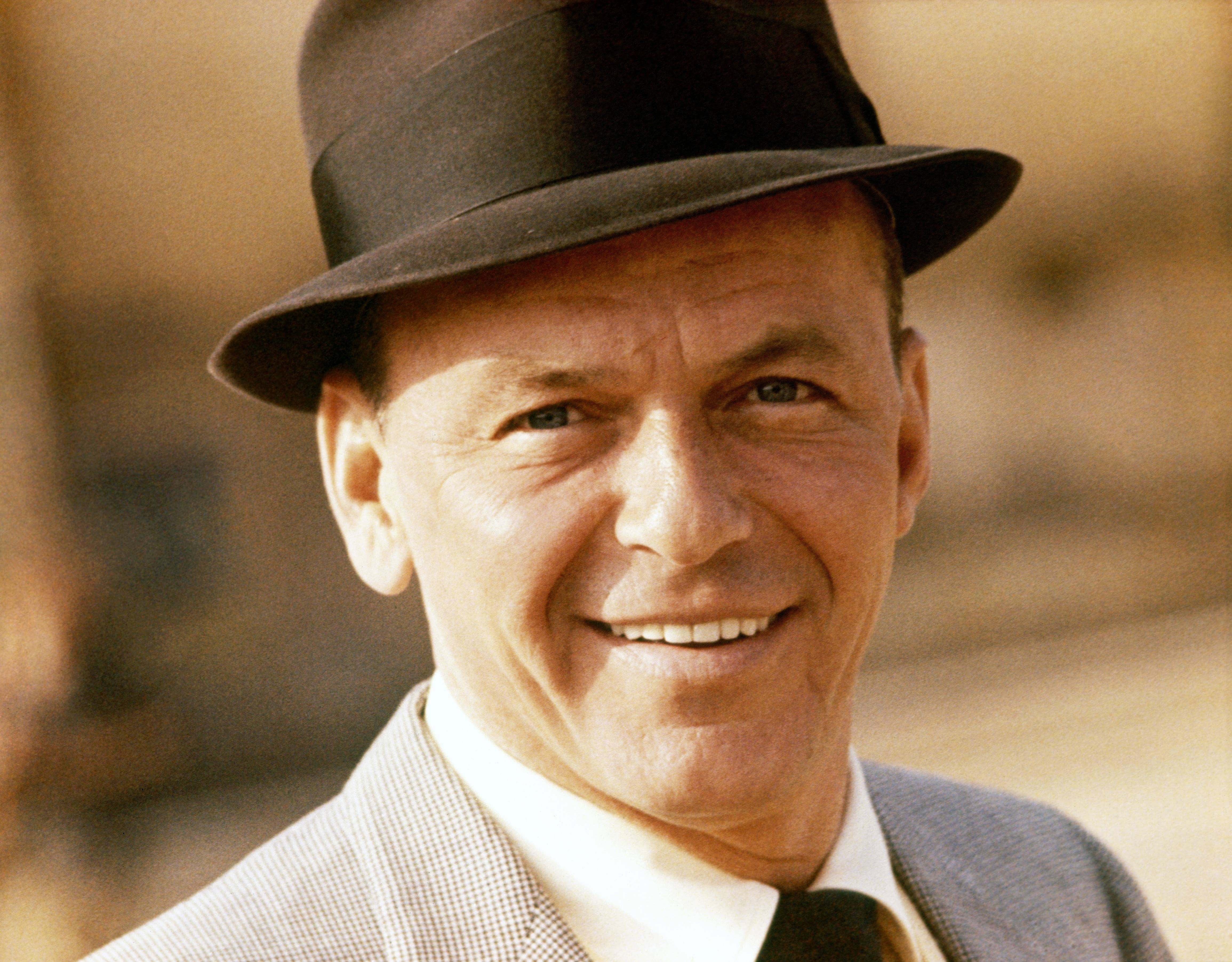 Sinatra 桌布 壁紙 — 一個戴著黑色費多拉帽和西裝外套的微笑男子，用溫暖的眼神直視鏡頭。