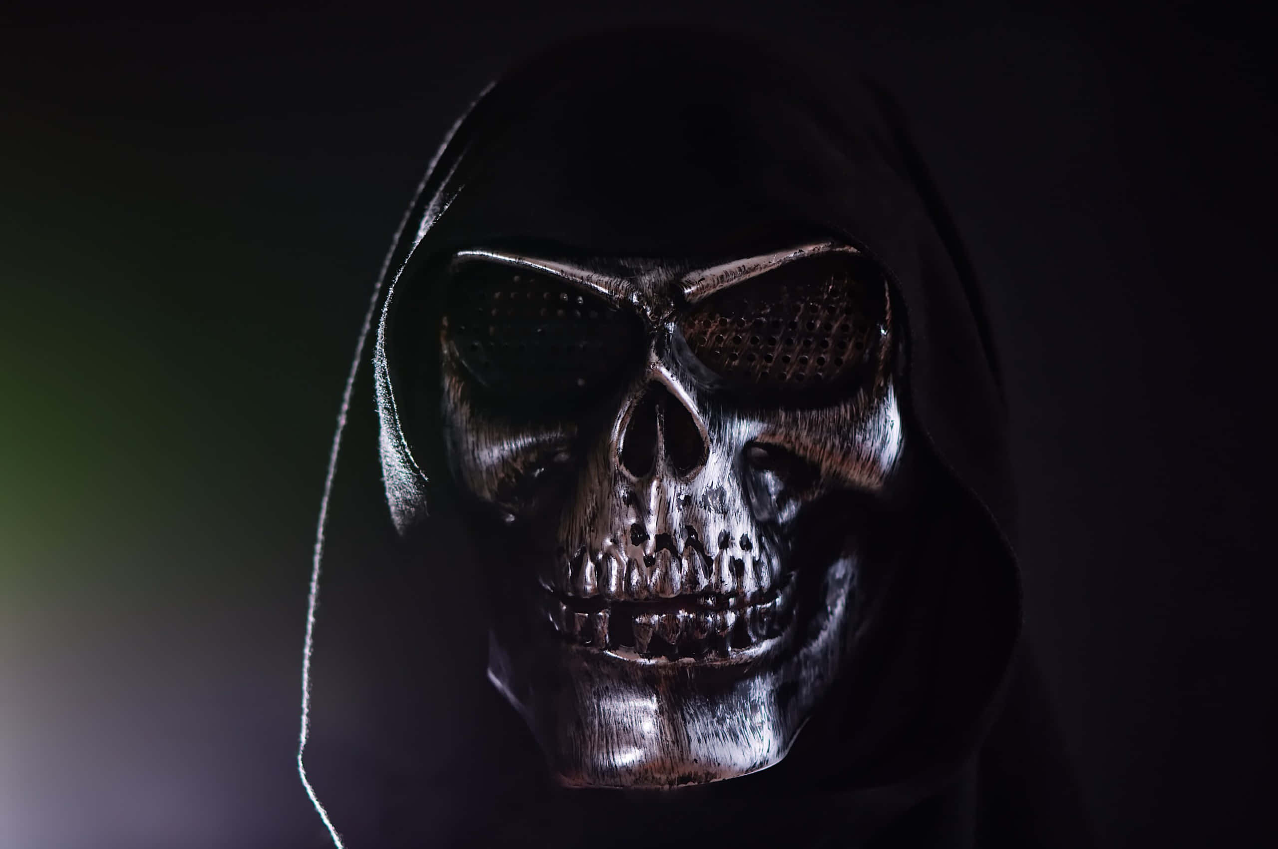 Horror Masks 桌布 壁紙 — 一個戴著黑色連帽衫、帶網眼眼罩的可怕骷髏臉人物，在黑暗背景下戲劇性的燈光下發光。