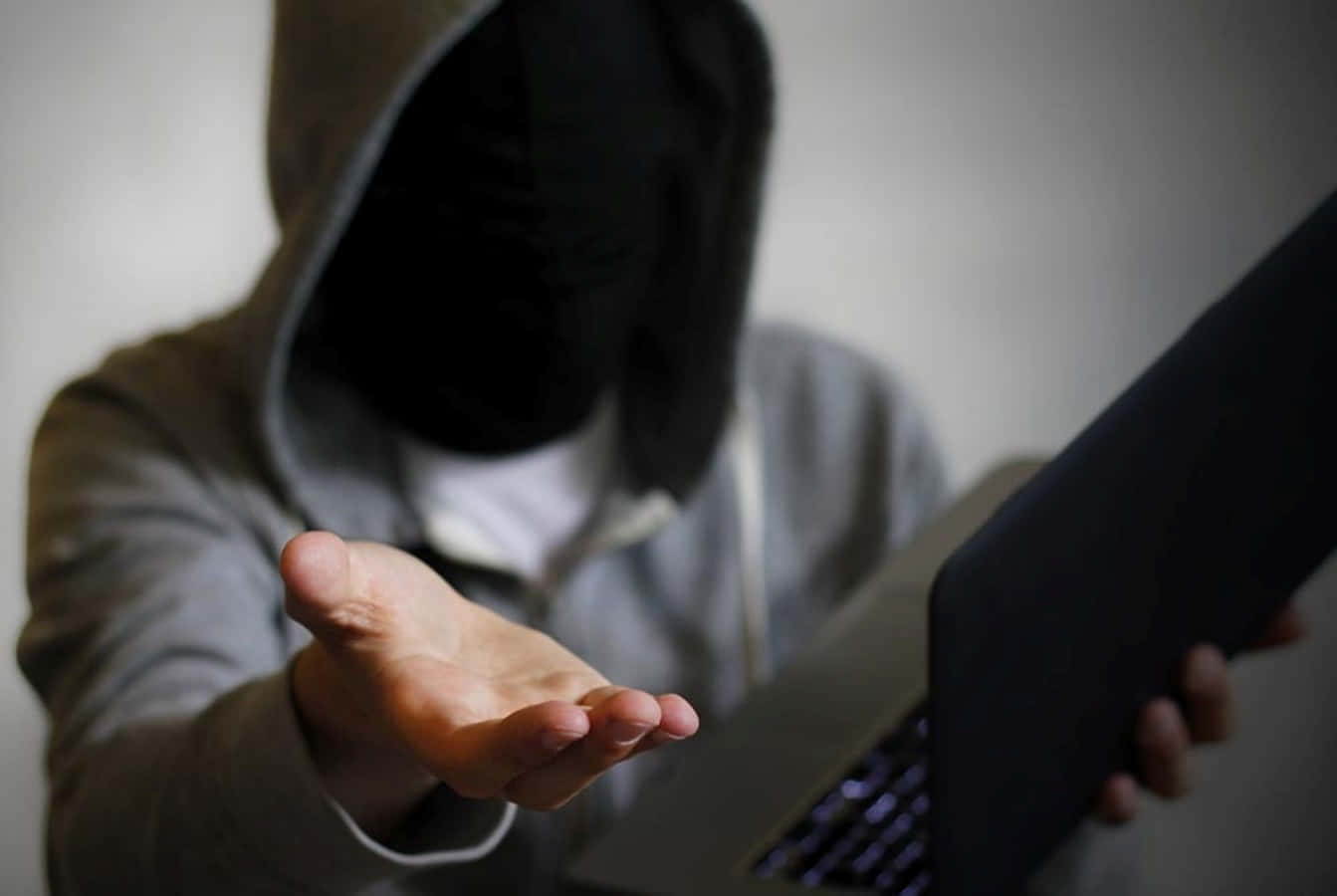 Blackmail 桌布 壁紙 — 一個戴著黑色面具的兜帽人物，在昏暗的房間裡拿著筆記型電腦，同時向觀看者伸出手。