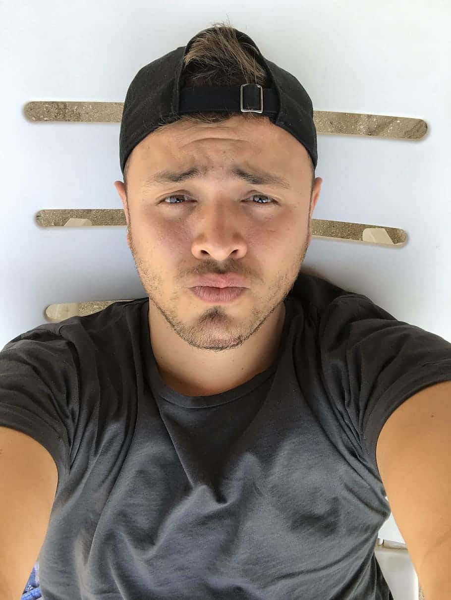 Selfie Man 桌布 壁紙 — 一個戴著黑色鴨舌帽和灰色T恤的年輕男子，背靠著有木條的白色牆壁躺著，直接看著鏡頭，表情俏皮。