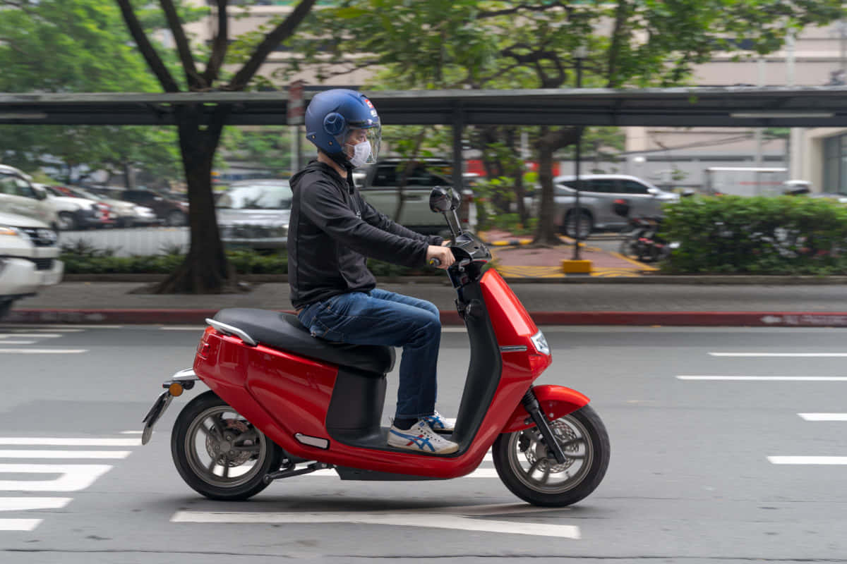 scooter 桌布 壁紙 — 一個戴藍色頭盔的人騎著一輛亮紅色的滑板車，穿過一條兩旁有綠色樹木的模糊城市街道。
