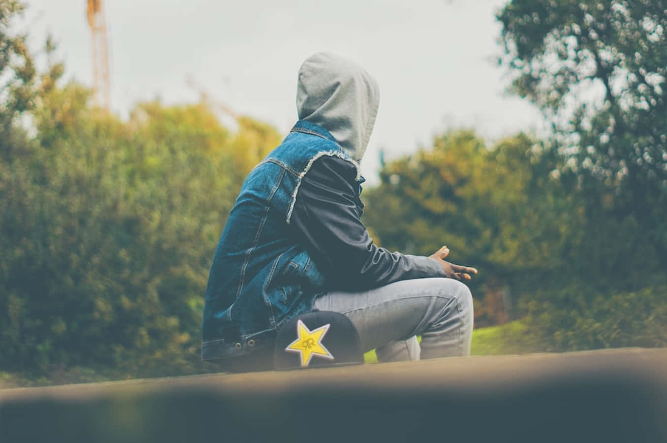 profile pic 桌布 壁紙 — 一個戴連帽衫、穿牛仔外套的人獨自坐在邊緣上，背景模糊中有樹木和起重機。
