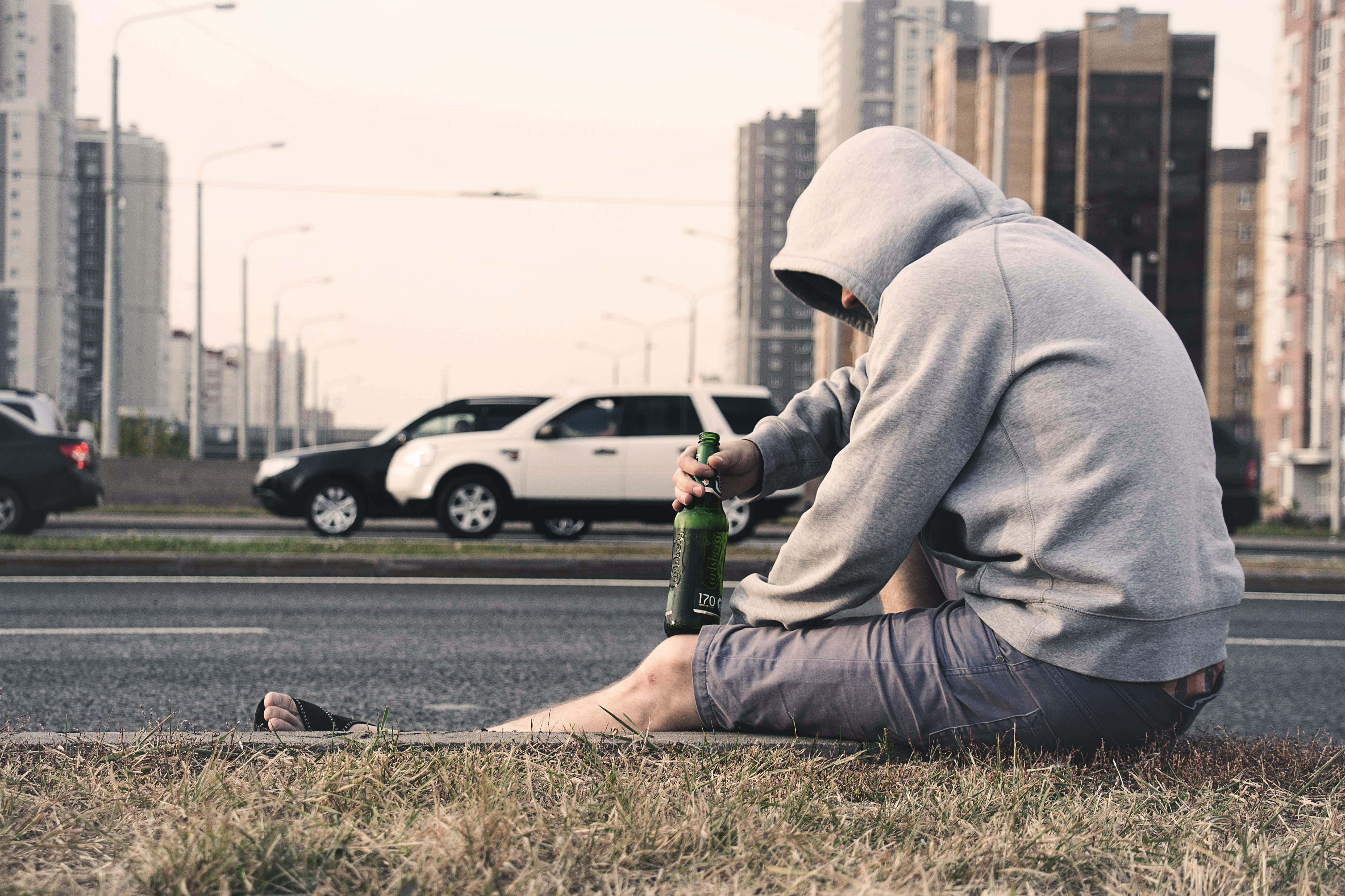 Drunk 桌布 壁紙 — 一個戴連帽衫的人獨自坐在路邊，手持啤酒瓶，汽車在都市景觀中駛過。
