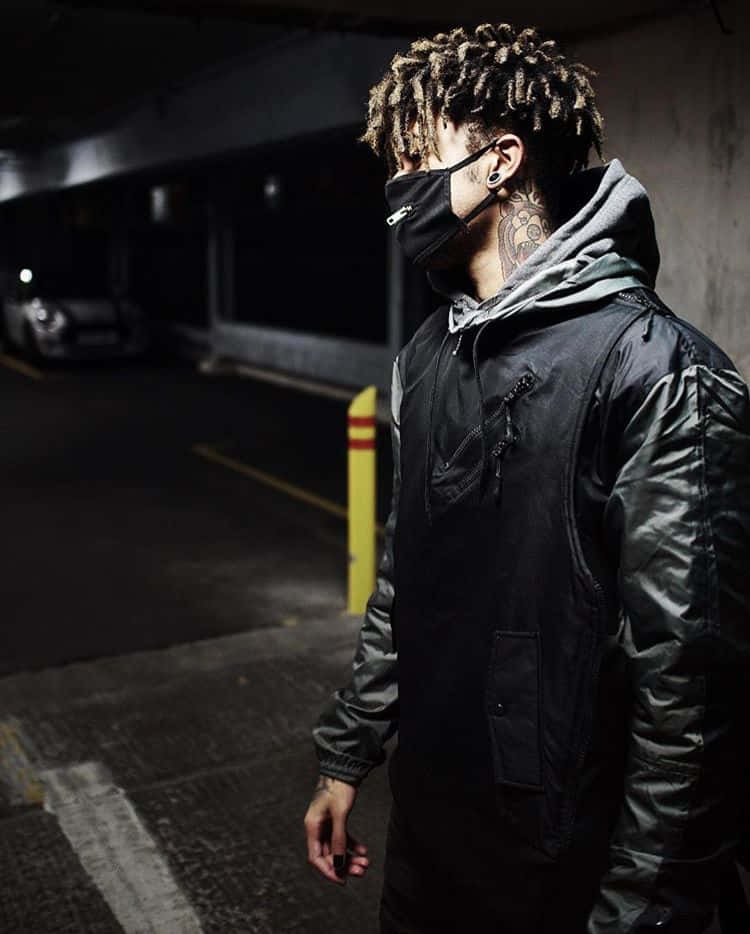 Mask Boy 桌布 壁紙 — 一個戴面具、有辮子頭和刺青的男人，身處燈光昏暗的地下停車場，穿著黑色外套。