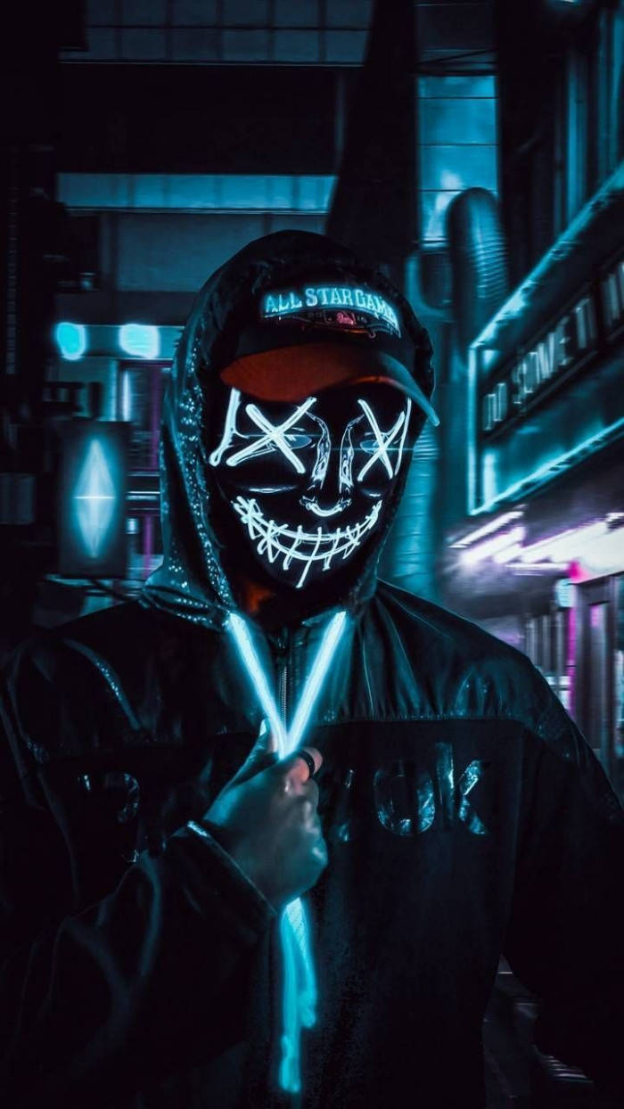 hacker 3d 桌布 壁紙 — 一個戴面具、穿連帽衫的人物在黑暗的城市後巷霓虹燈下拿著發光的棒子。