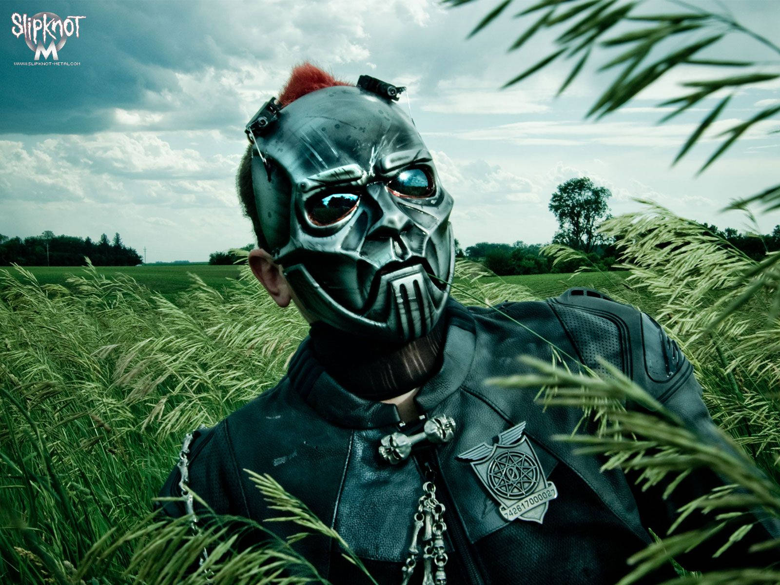 Slipknot 桌布 壁紙 — 一個戴面具、紅色莫西干頭的人站在高高的綠草中，天空多雲。