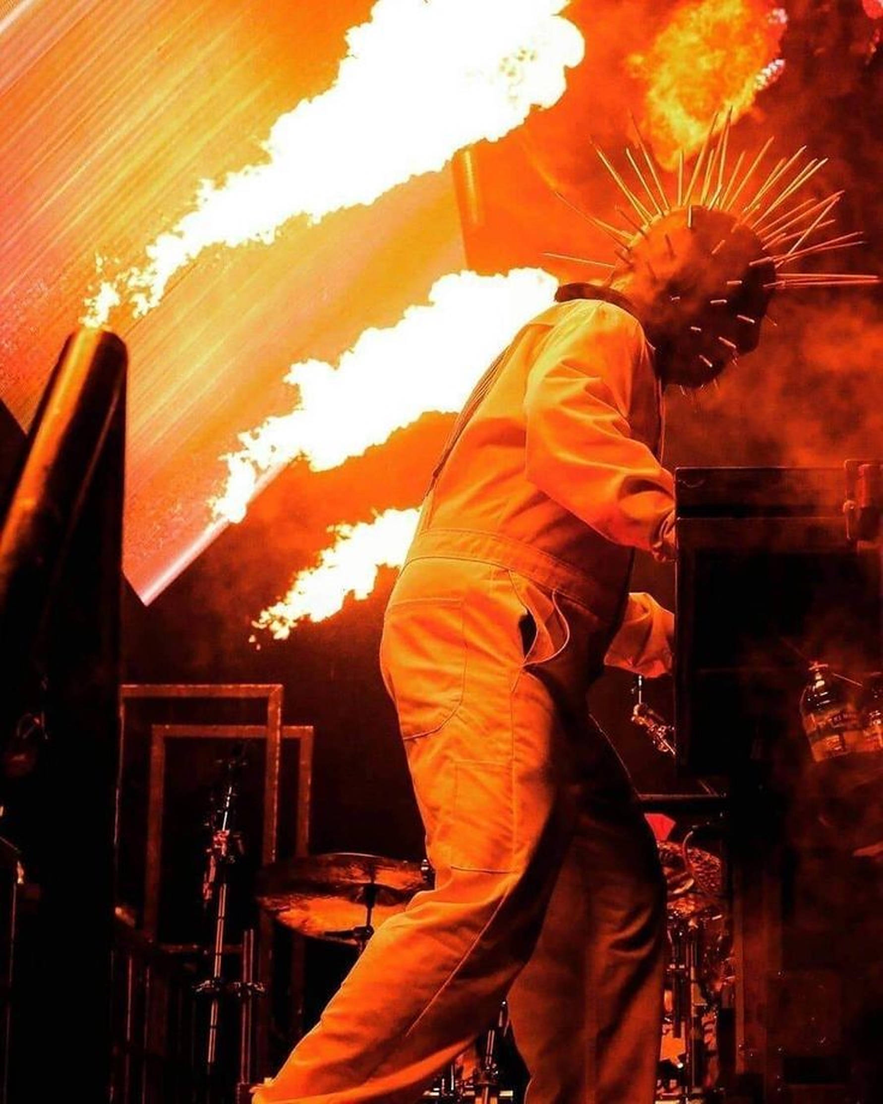 craig jones 桌布 壁紙 — 一個戴面具、身穿橙色連身衣的人物在黑暗舞台上打鼓，身後爆發熾熱爆炸。