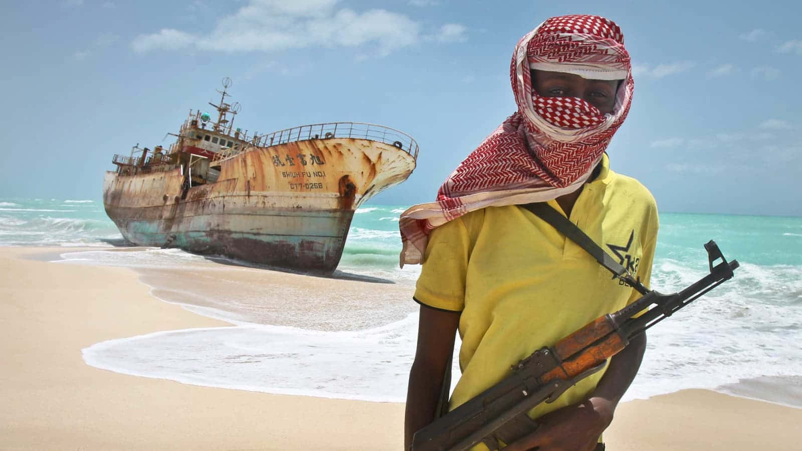 Pirate 桌布 壁紙 — 一個戴面具的武裝男子穿著黃色襯衫，站在沙灘上，旁邊是生鏽的沉船，背景是明亮的藍天。