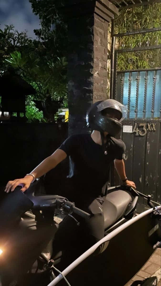情侶 桌布 壁紙 — 一個戴頭盔的人夜晚坐在一輛時尚的黑色摩托車上，旁邊是一個有閘門的入口，周圍有茂密的綠色植物。