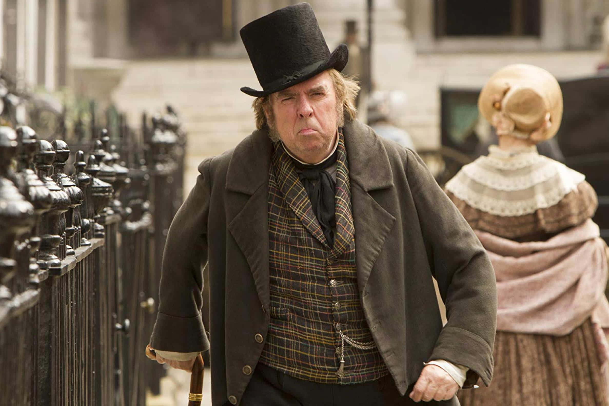 Timothy Spall 桌布 壁紙 — 一個戴高帽、穿格紋背心的暴躁男人，身後跟著一個戴帽子的女人，走在鵝卵石街上。