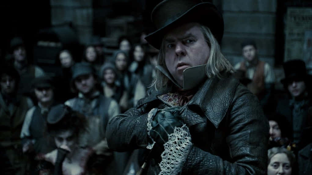 Timothy Spall 桌布 壁紙 — 一個戴高帽、穿皮外套的男人站在昏暗、骯髒的城市環境中，在人群面前的舞台中央。