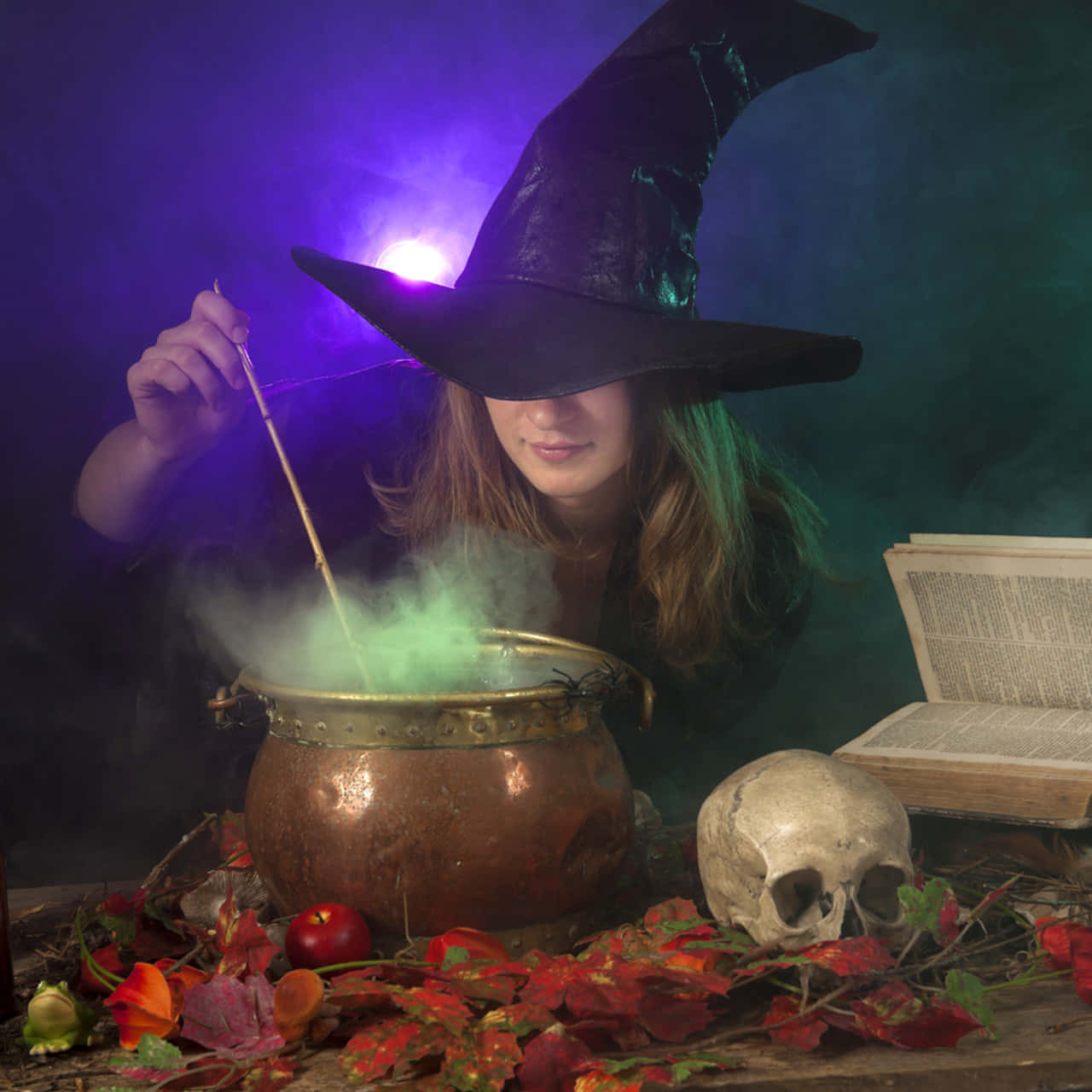 Witching Hour 桌布 壁紙 — 一個戴黑帽的女巫在銅鍋中攪拌發光的綠色藥水，周圍有秋葉和一個頭骨。