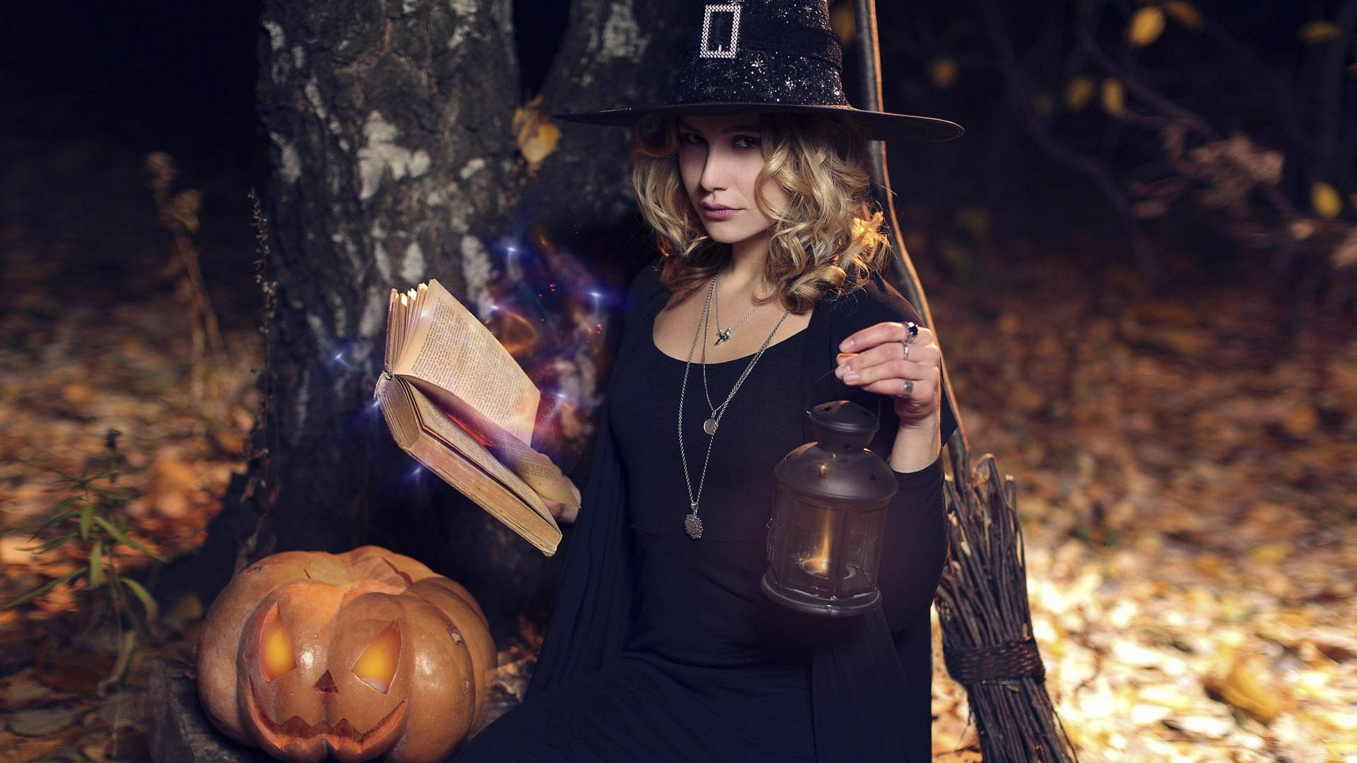 Witchy 桌布 壁紙 — 一個戴黑帽穿黑斗篷的女巫坐在森林裡，拿著發光的書和燈籠，旁邊有一個雕刻的南瓜。
