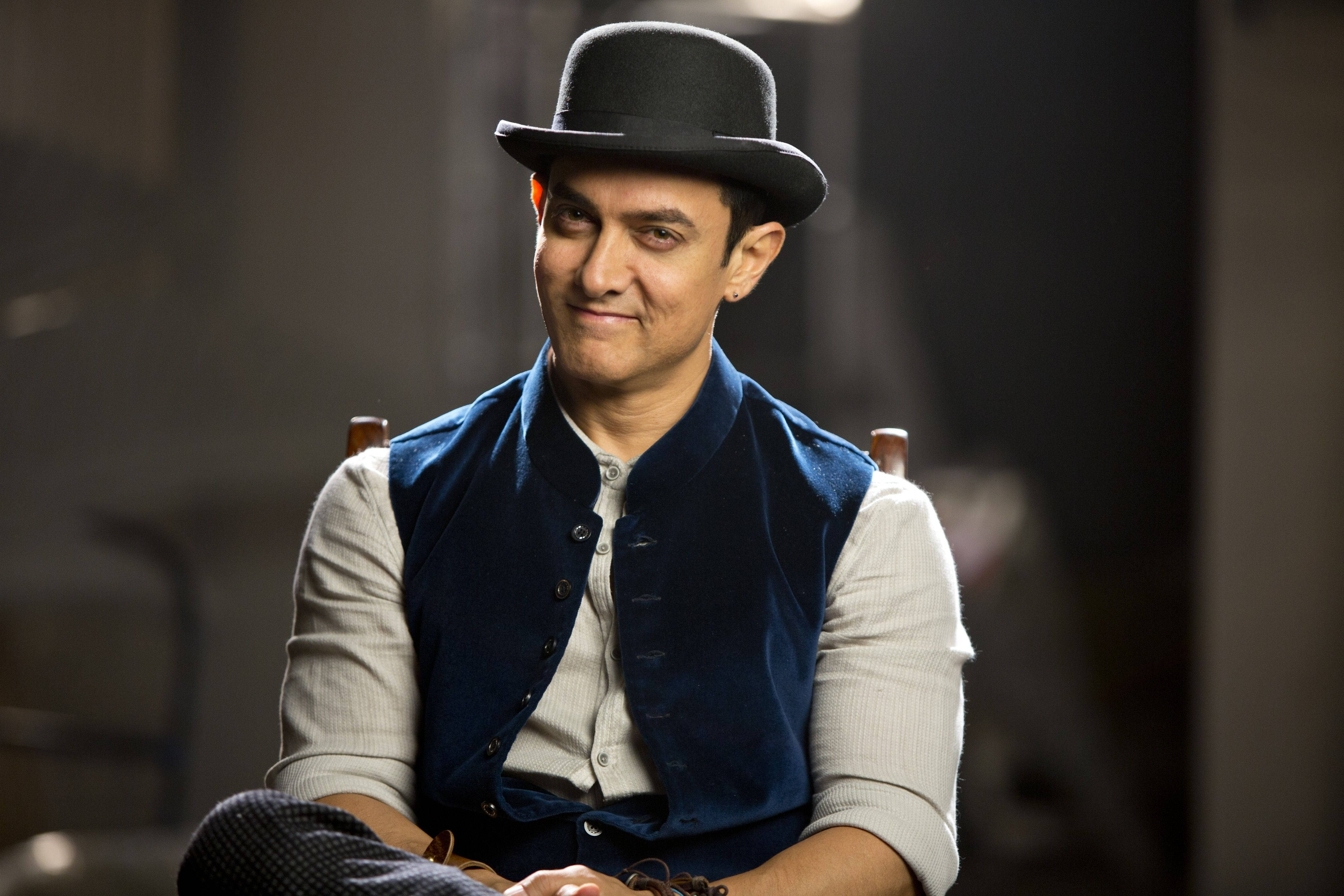 Aamir Khan 桌布 壁紙 — 一個戴黑色圓頂禮帽、穿藍色背心的男人坐在昏暗的房間裡，露出微妙的笑意。