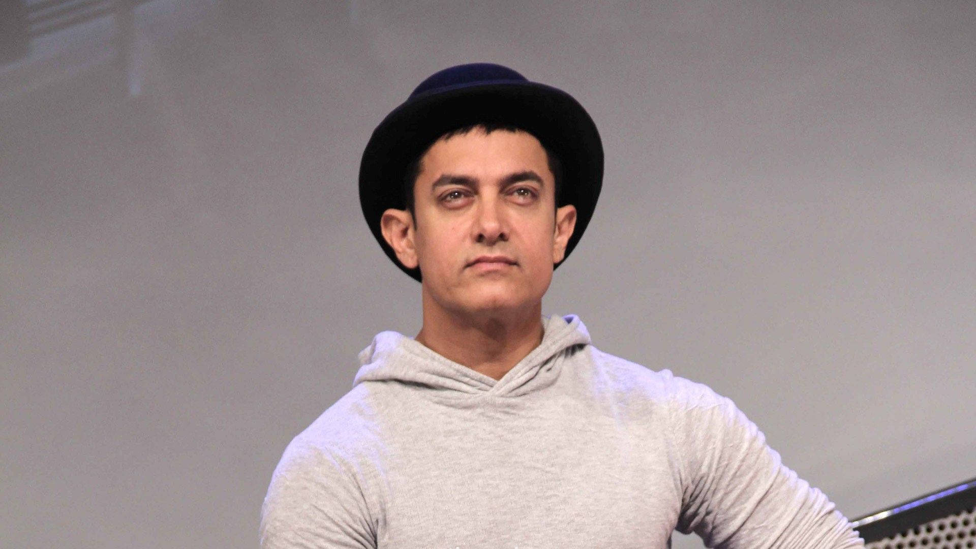 Aamir Khan 桌布 壁紙 — 一個戴黑色圓頂禮帽和灰色連帽衫的男人，在純灰色背景前表情嚴肅地站著。