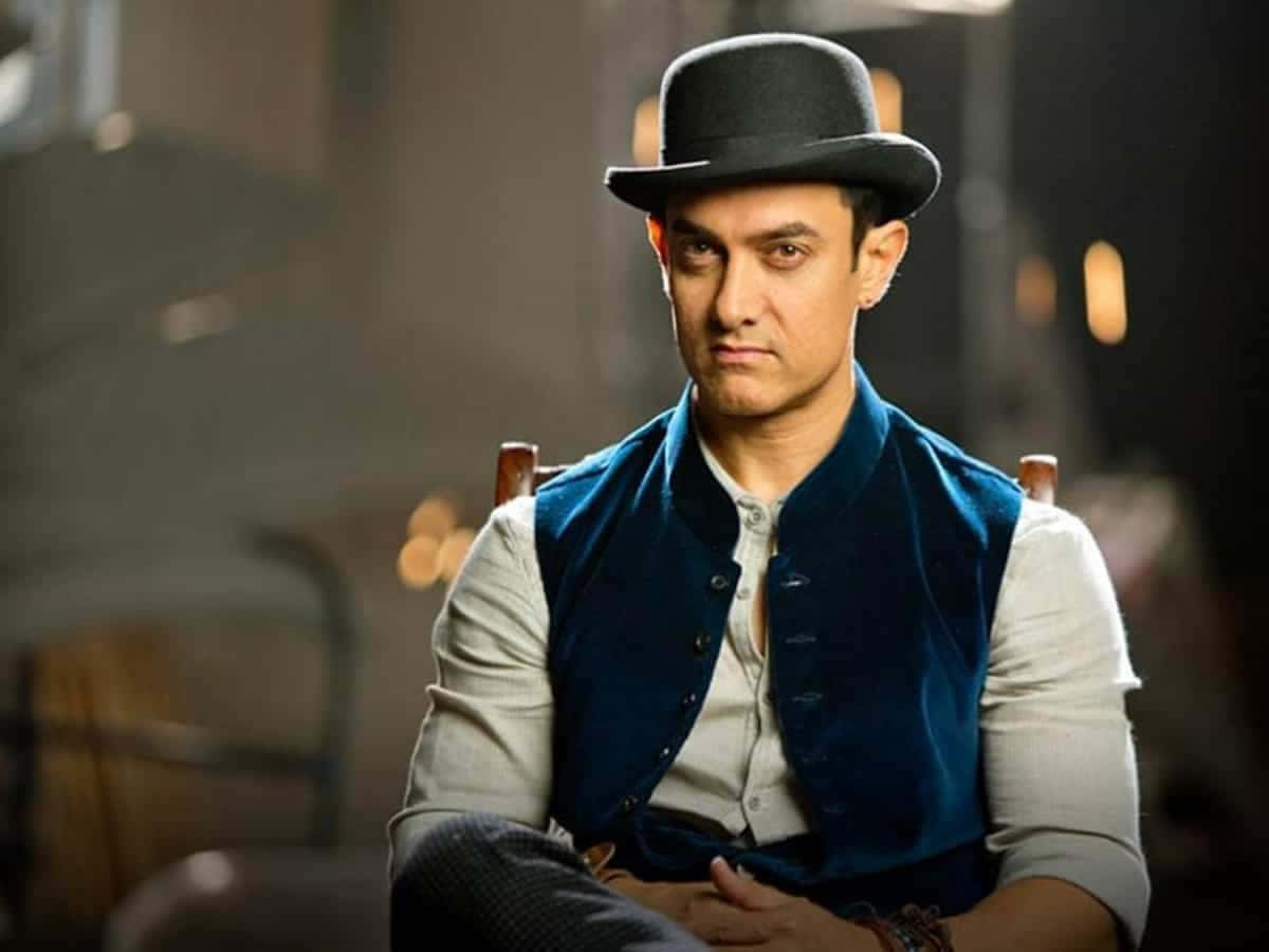 Aamir Khan 桌布 壁紙 — 一個戴黑色圓頂禮帽和藍色天鵝絨背心的男人，在燈光昏暗的房間裡表情嚴肅地坐著。