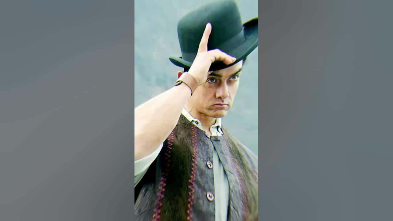 Aamir Khan 桌布 壁紙 — 一個戴黑色圓頂禮帽的男人用手指將帽子傾斜，身穿毛邊背心，背景為柔和的灰色。