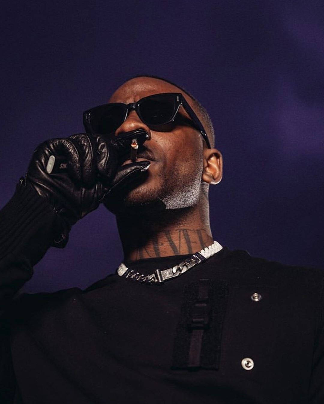 Skepta 桌布 壁紙 — 一個戴黑色太陽眼鏡和皮手套的男人在深紫色背景前抽大麻煙。