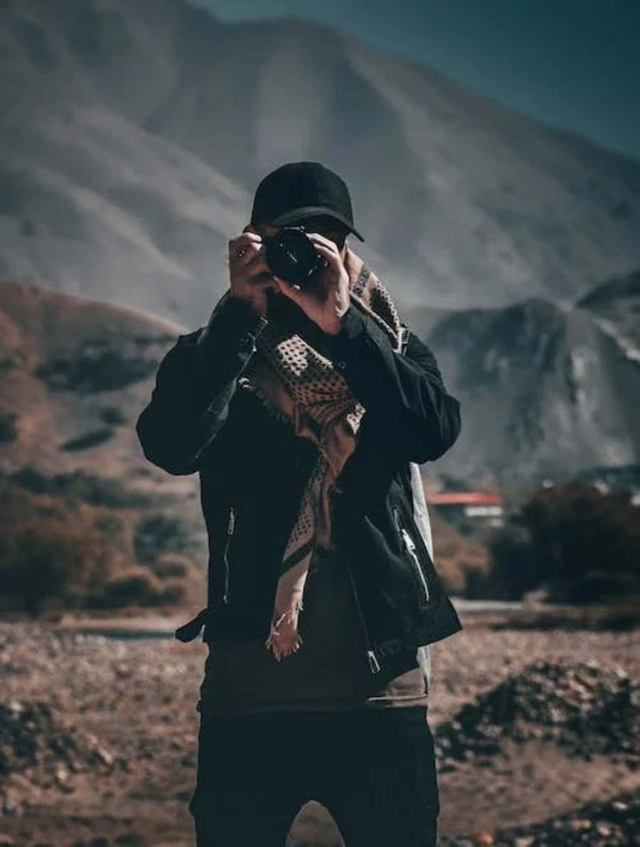profile pic 桌布 壁紙 — 一個戴黑色帽子、穿黑色外套的人，在崎嶇的山景背景下將相機舉到面前。