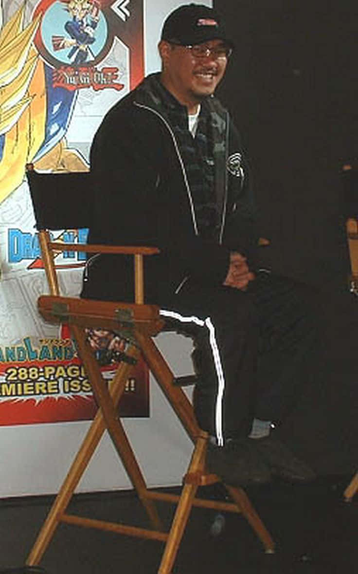 Akira Toriyama 桌布 壁紙 — 一個戴黑色帽子、穿黑色外套的微笑男人坐在木製導演椅上，前面有一張七龍珠海報。