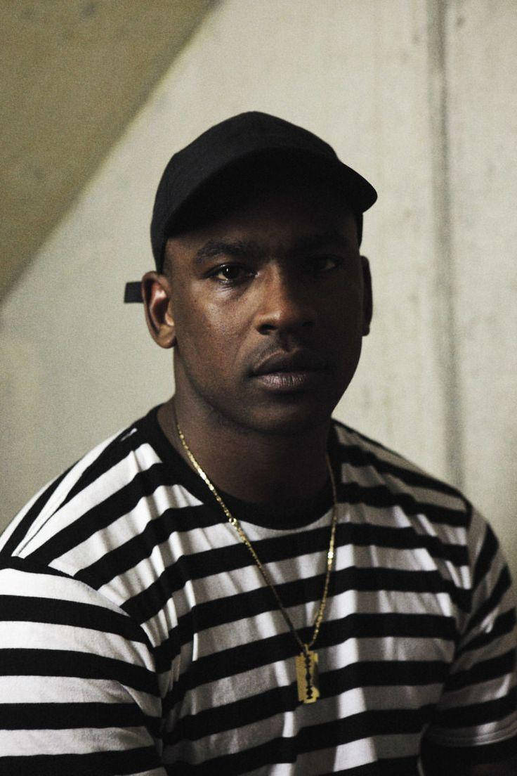 Skepta 桌布 壁紙 — 一個戴黑色帽子、身穿黑白條紋襯衫的男人，對著一面普通的混凝土牆凝視。