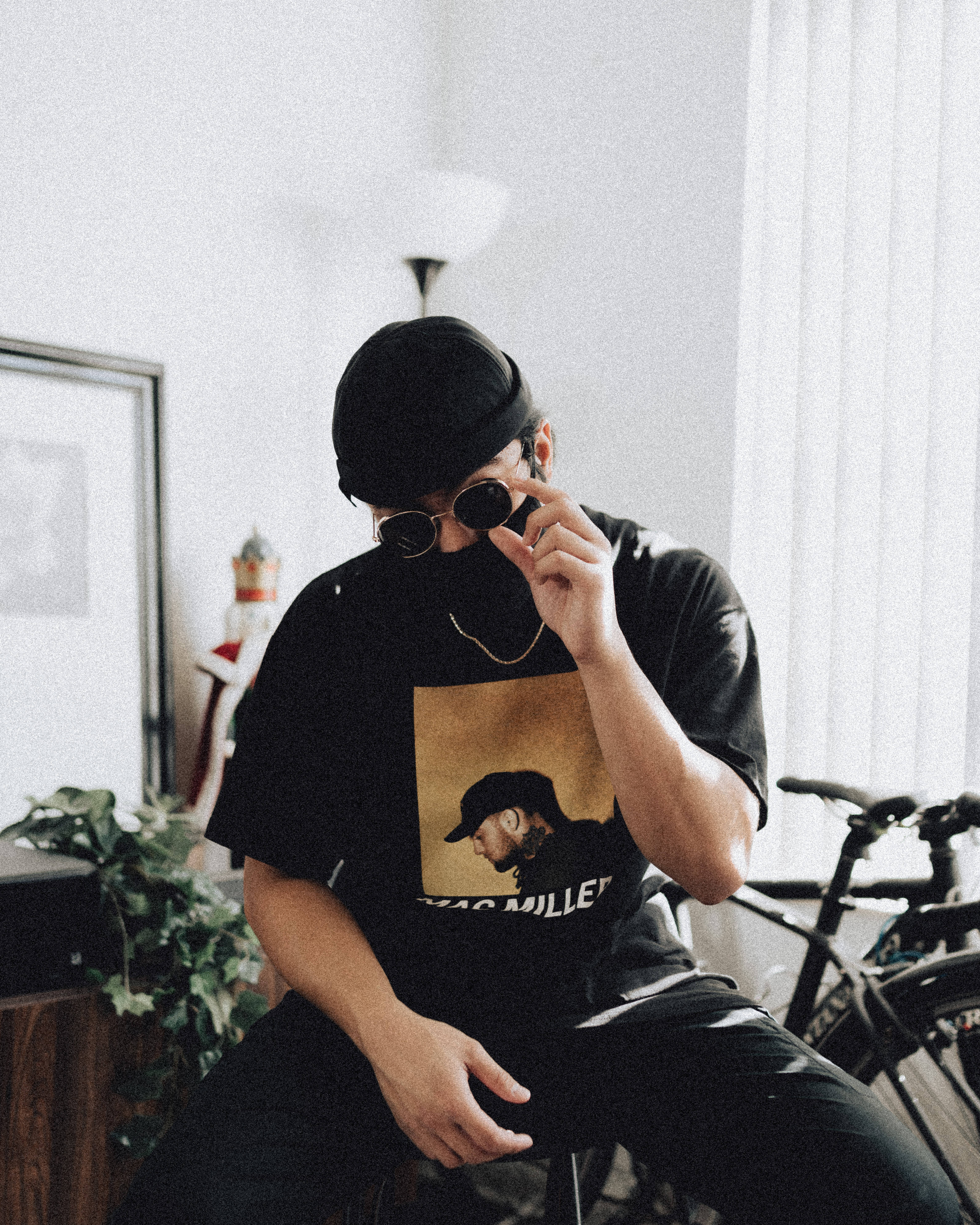 Mac Miller 桌布 壁紙 — 一個戴黑色帽子和太陽眼鏡的人坐在室內靠近自行車和盆栽的地方調整眼鏡。