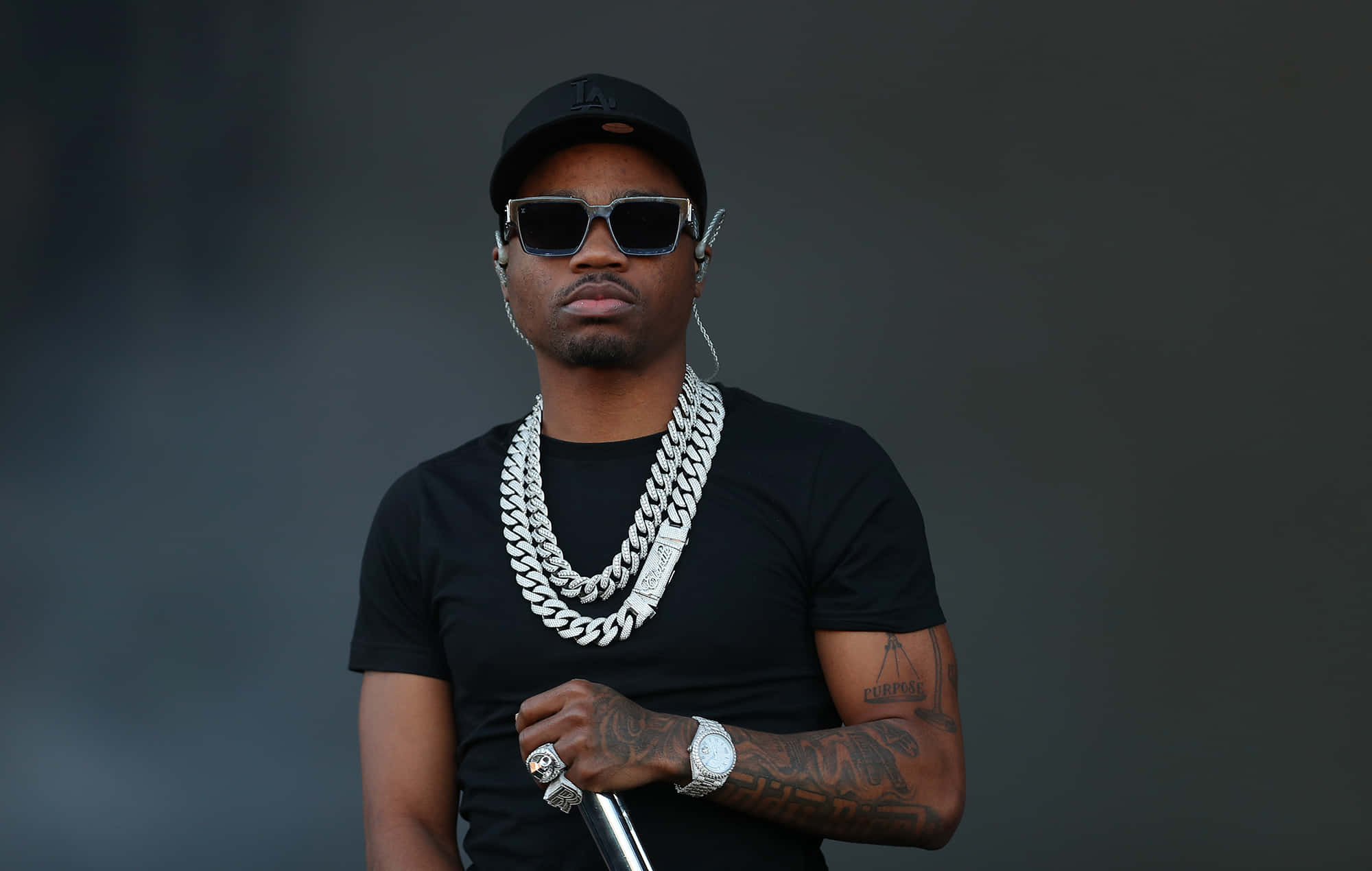 Roddy Ricch 桌布 壁紙 — 一個戴黑色帽子和太陽眼鏡的男人，戴著一條粗銀鏈，在深灰色背景前拿著麥克風。