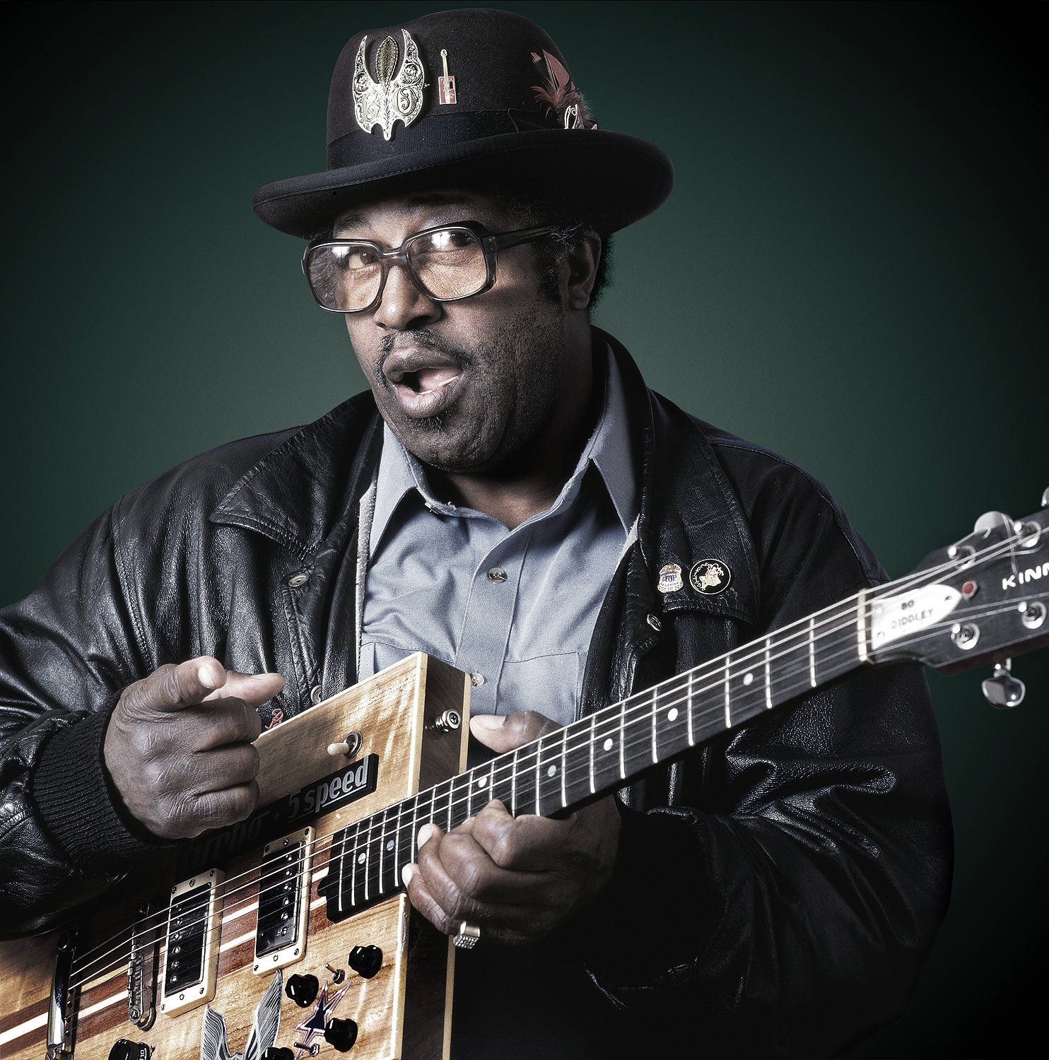 Bo Diddley 桌布 壁紙 — 一個戴黑色帽子和眼鏡的男人在深綠色背景前熱情地彈奏木製電吉他。