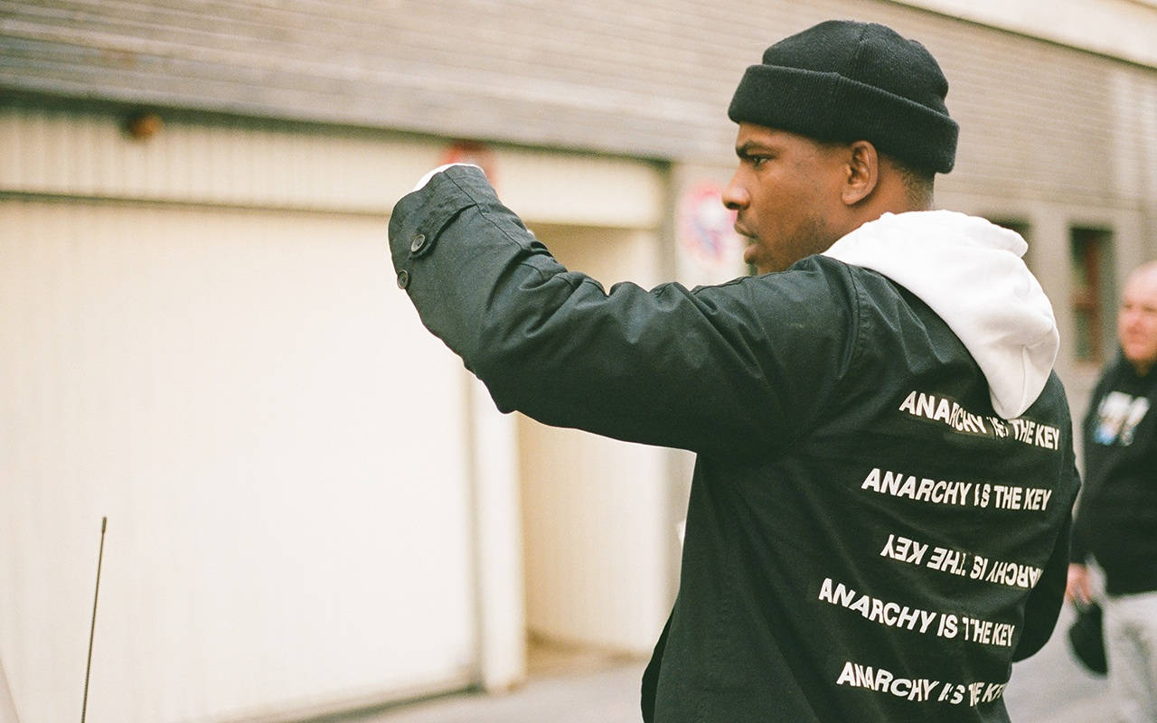 Skepta 桌布 壁紙 — 一個戴黑色毛帽、穿夾克的男人，夾克背面印有「ANARCHY IS THE KEY」字樣，在都市街道上向前做手勢。