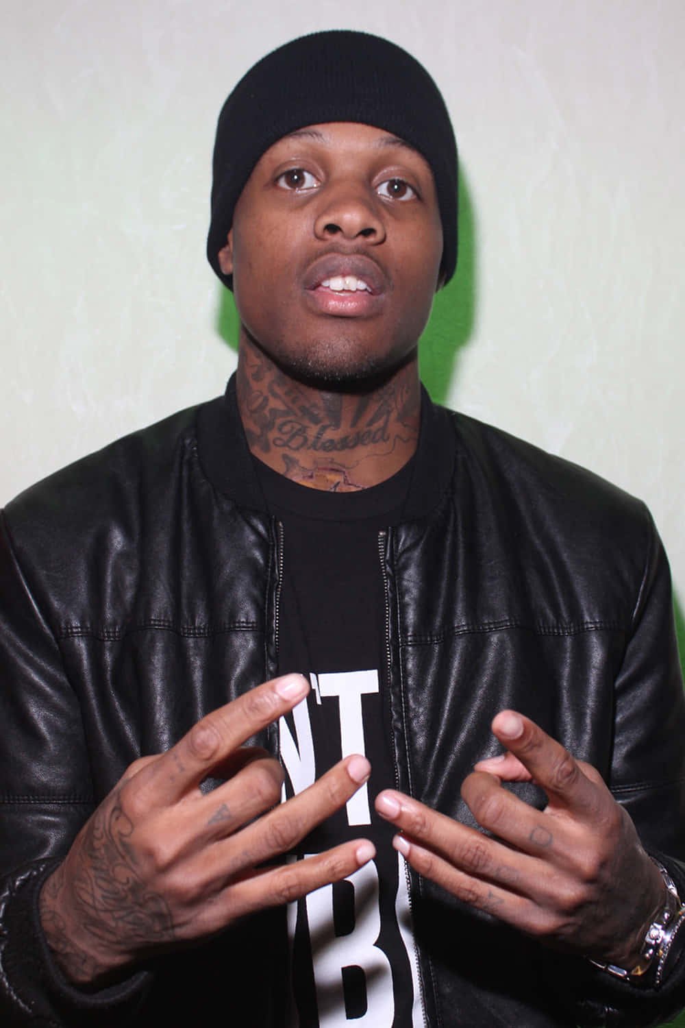 Lil Durk 桌布 壁紙 — 一個戴黑色毛帽、穿皮夾克的男人，在純綠色牆前說話時做手勢。