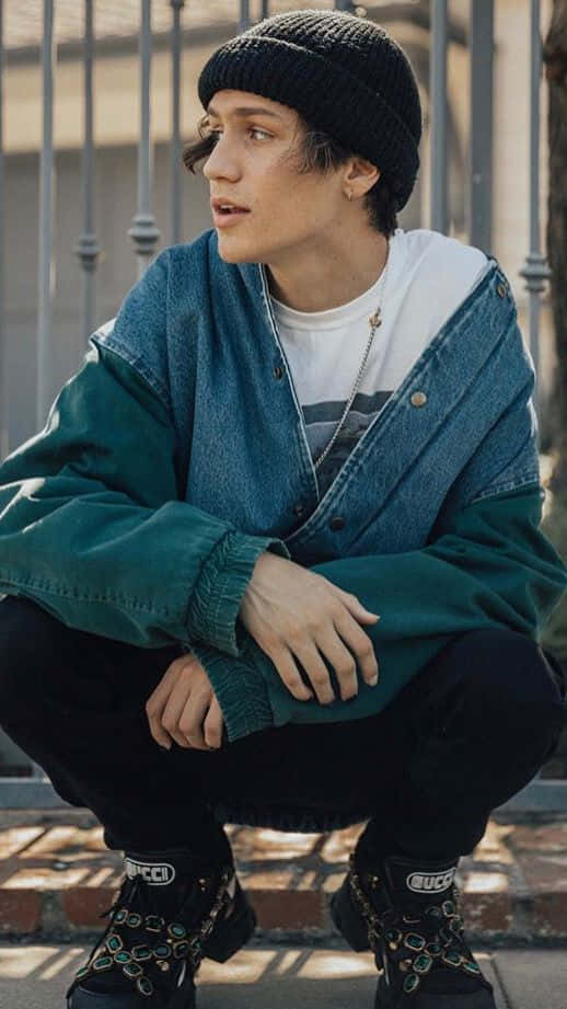 Denim 桌布 壁紙 — 一個戴黑色毛帽、身穿超大牛仔外套的年輕人，在金屬圍欄附近的磚人行道上蹲著。