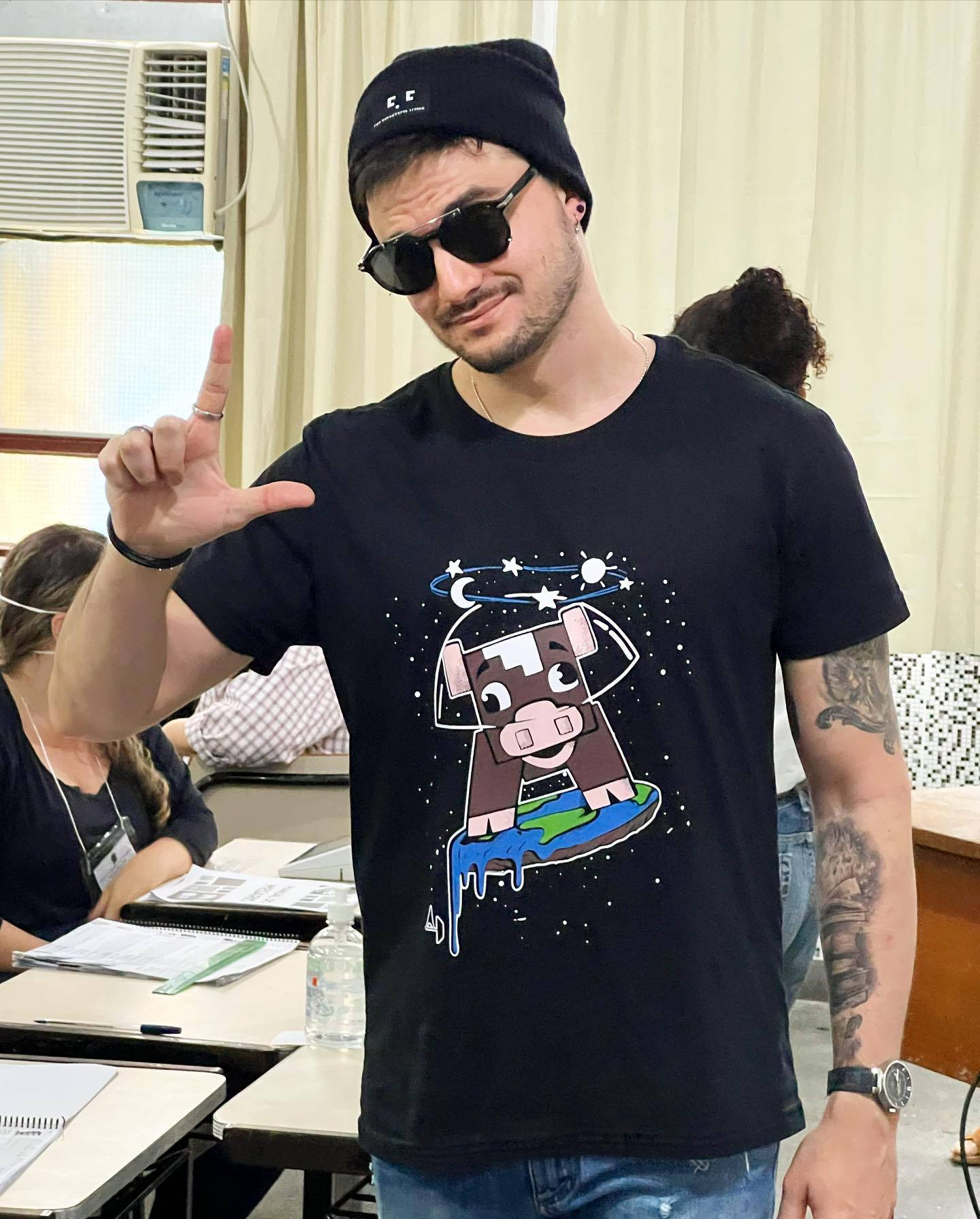 Mens Fashion 桌布 壁紙 — 一個戴黑色毛帽和太陽眼鏡的男人，穿著印有卡通牛在太空漂浮的T恤，比出和平手勢。