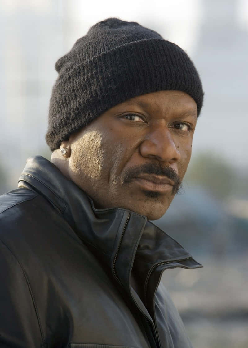 Ving Rhames 桌布 壁紙 — 一個戴黑色毛帽和皮夾克的男人，在模糊的城市背景下，神情嚴肅地凝視著。