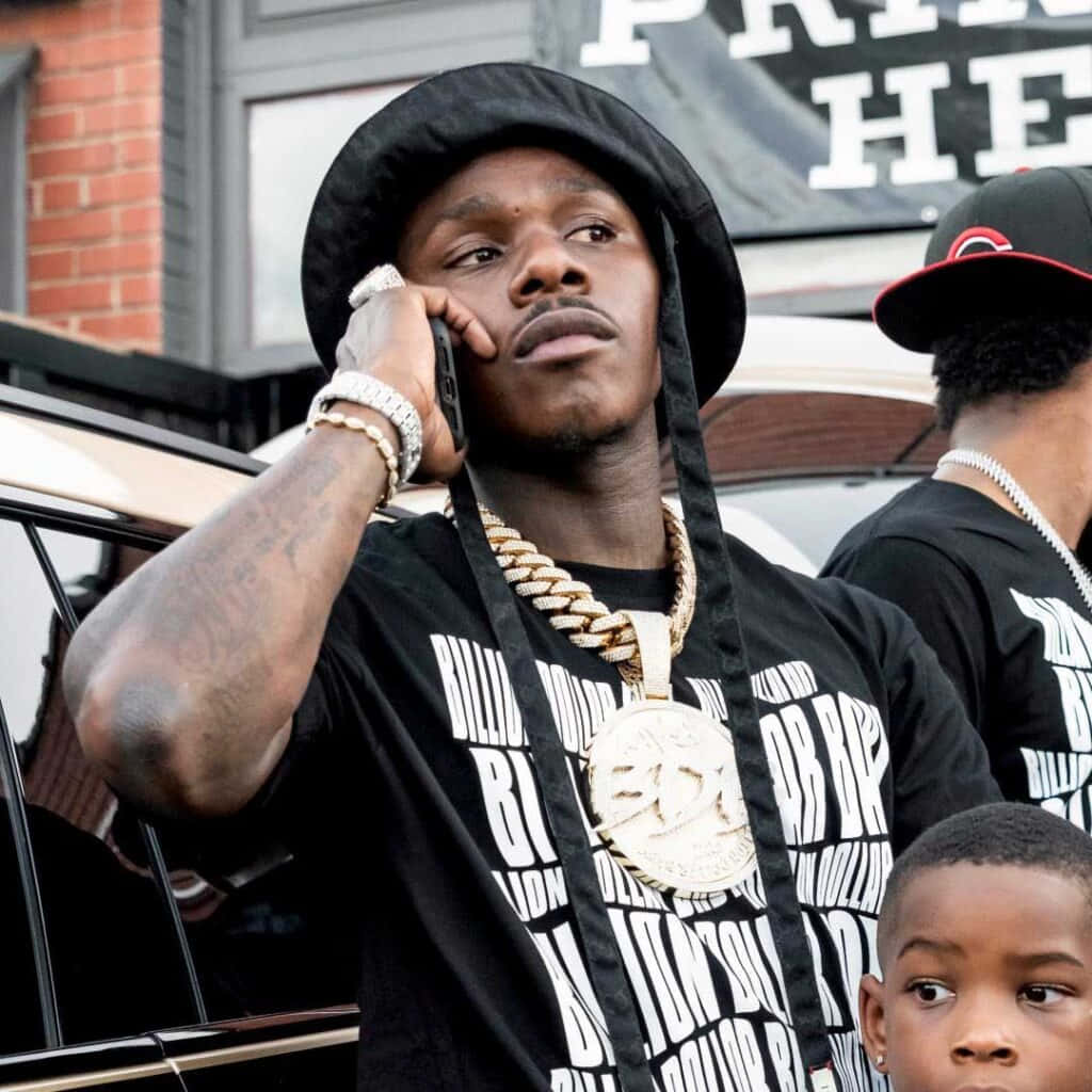Dababy 桌布 壁紙 — 一個戴黑色漁夫帽和金色鏈條的男人站在車旁，附近有一個小孩，正在講電話。