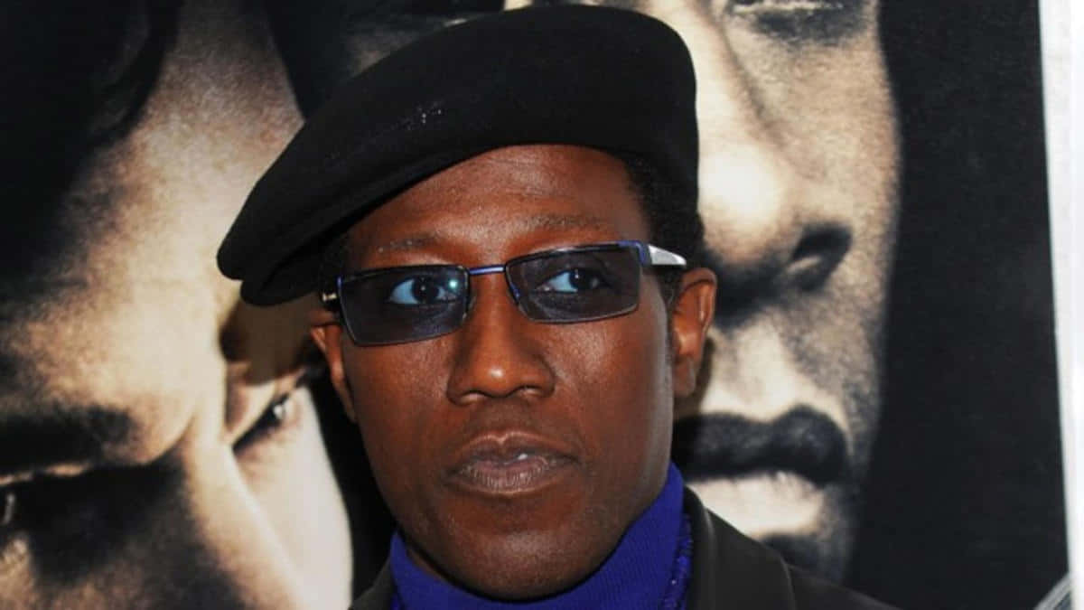 Wesley Snipes 桌布 壁紙 — 一個戴黑色貝雷帽和太陽眼鏡的男人站在一張臉部模糊的電影海報前。