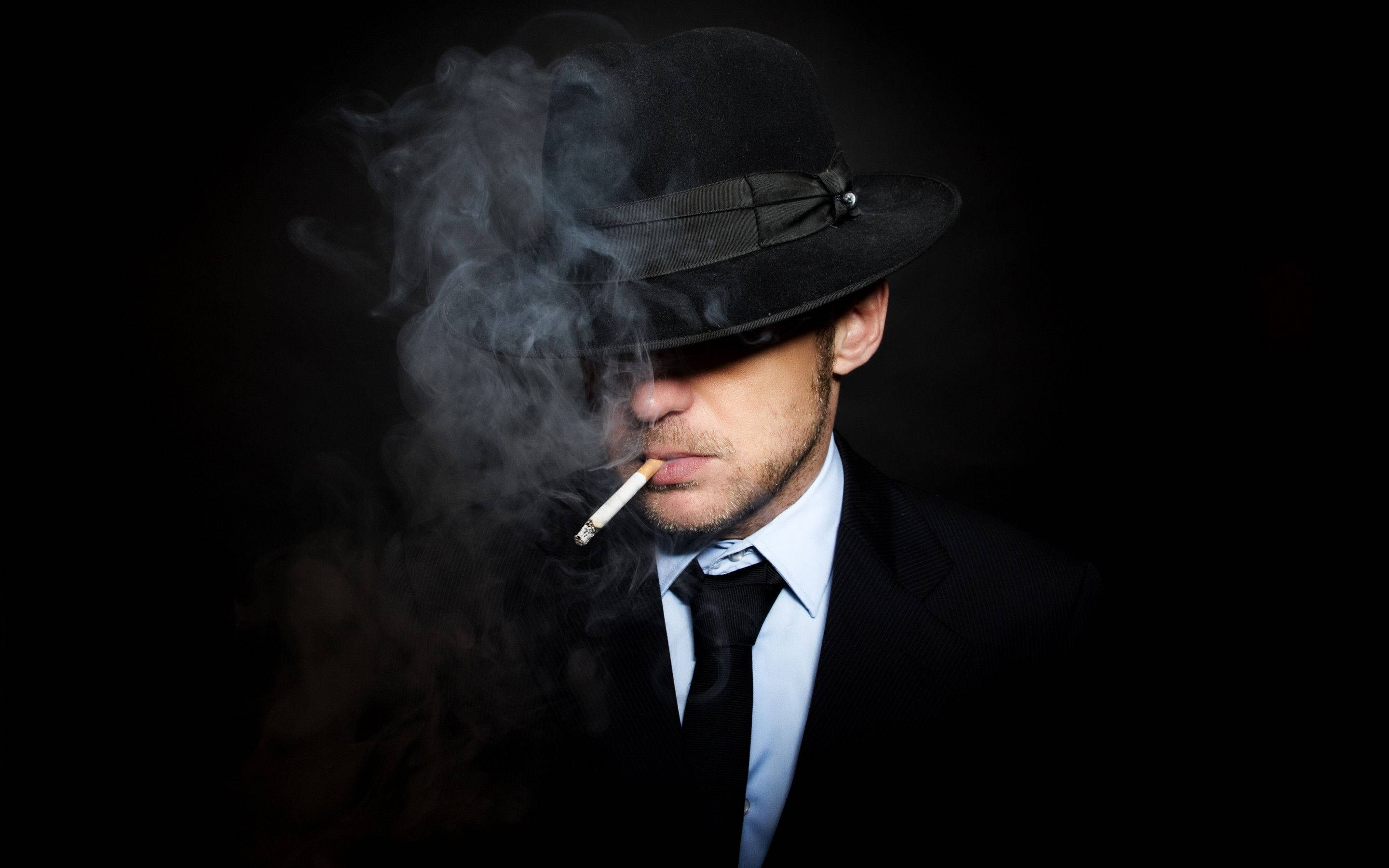 Man In Suit 桌布 壁紙 — 一個戴黑色軟呢帽、穿西裝的男人在黑暗背景下抽煙，煙霧繚繞。